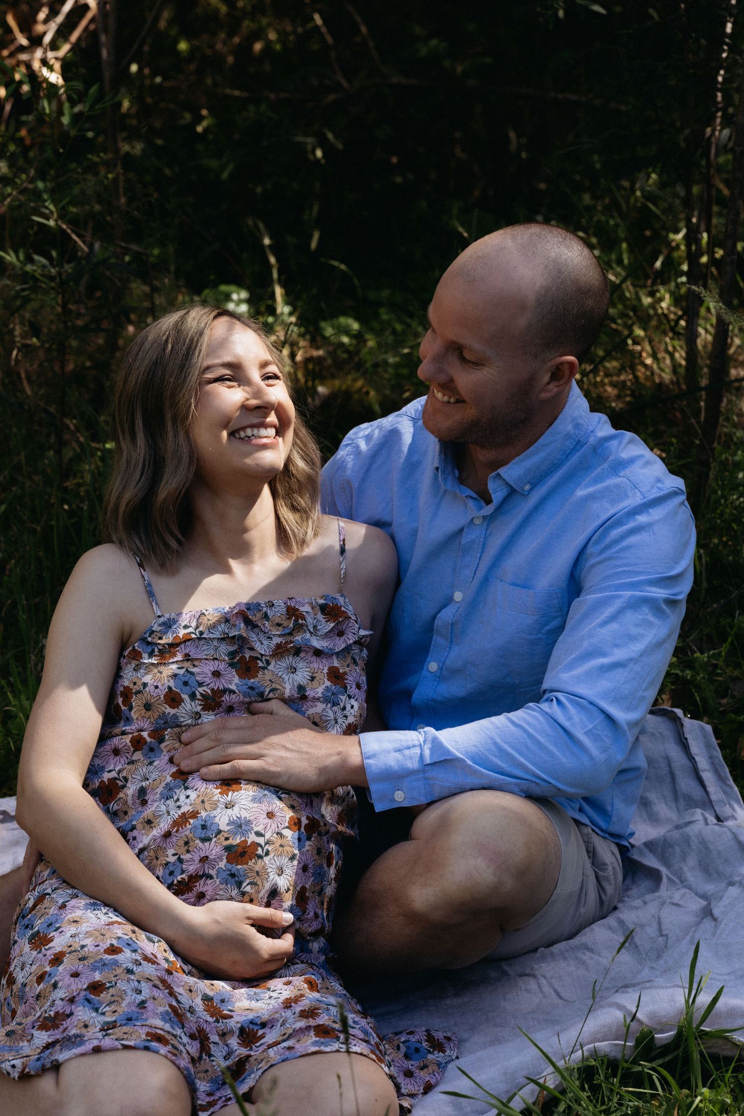 Outdoor maternity photography session featuring a smiling pregnant woman and her partner sitting on a blanket in a forest.