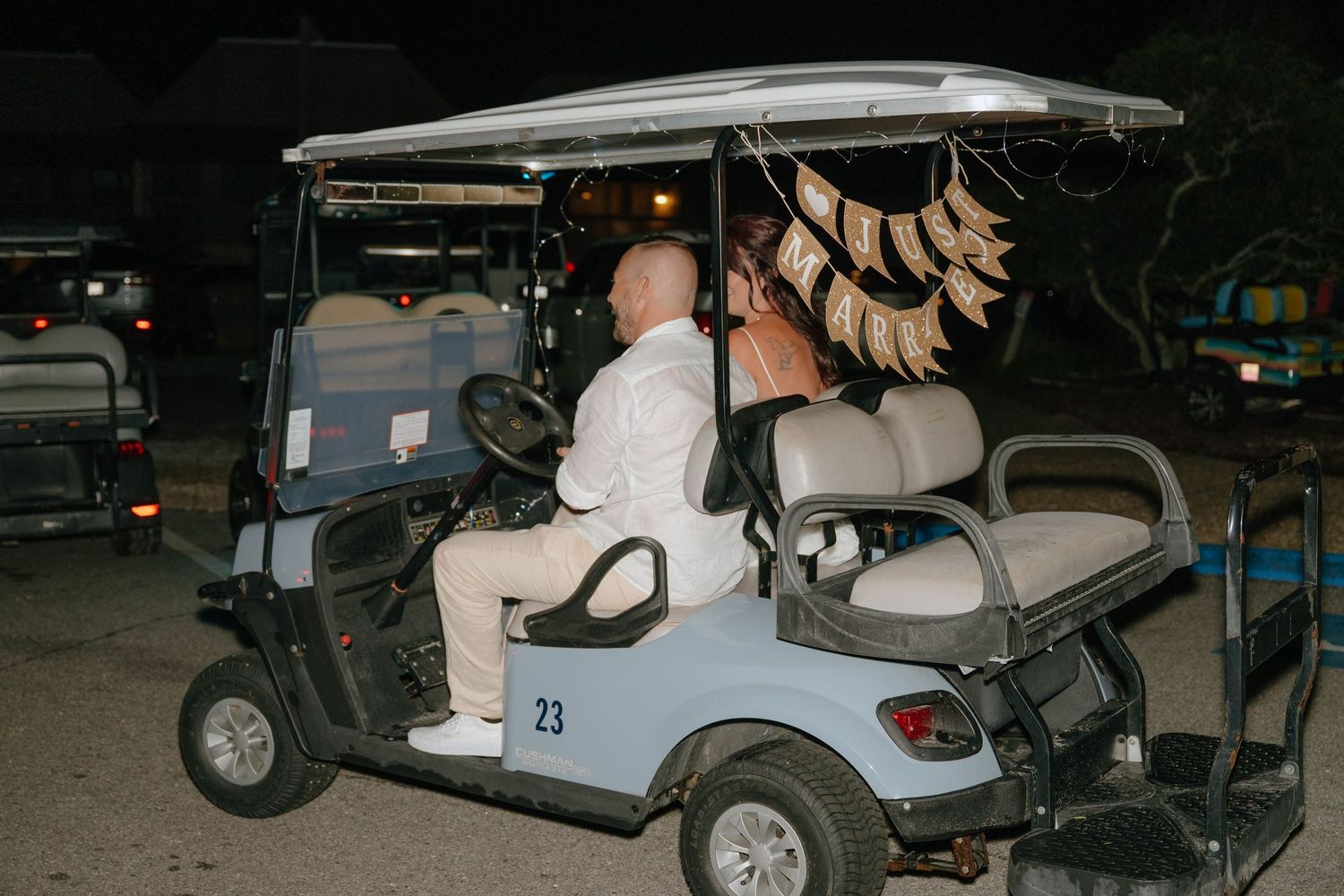 Groom in white outfit drives a decorated golf cart numbered 23 at night after the wedding reception.