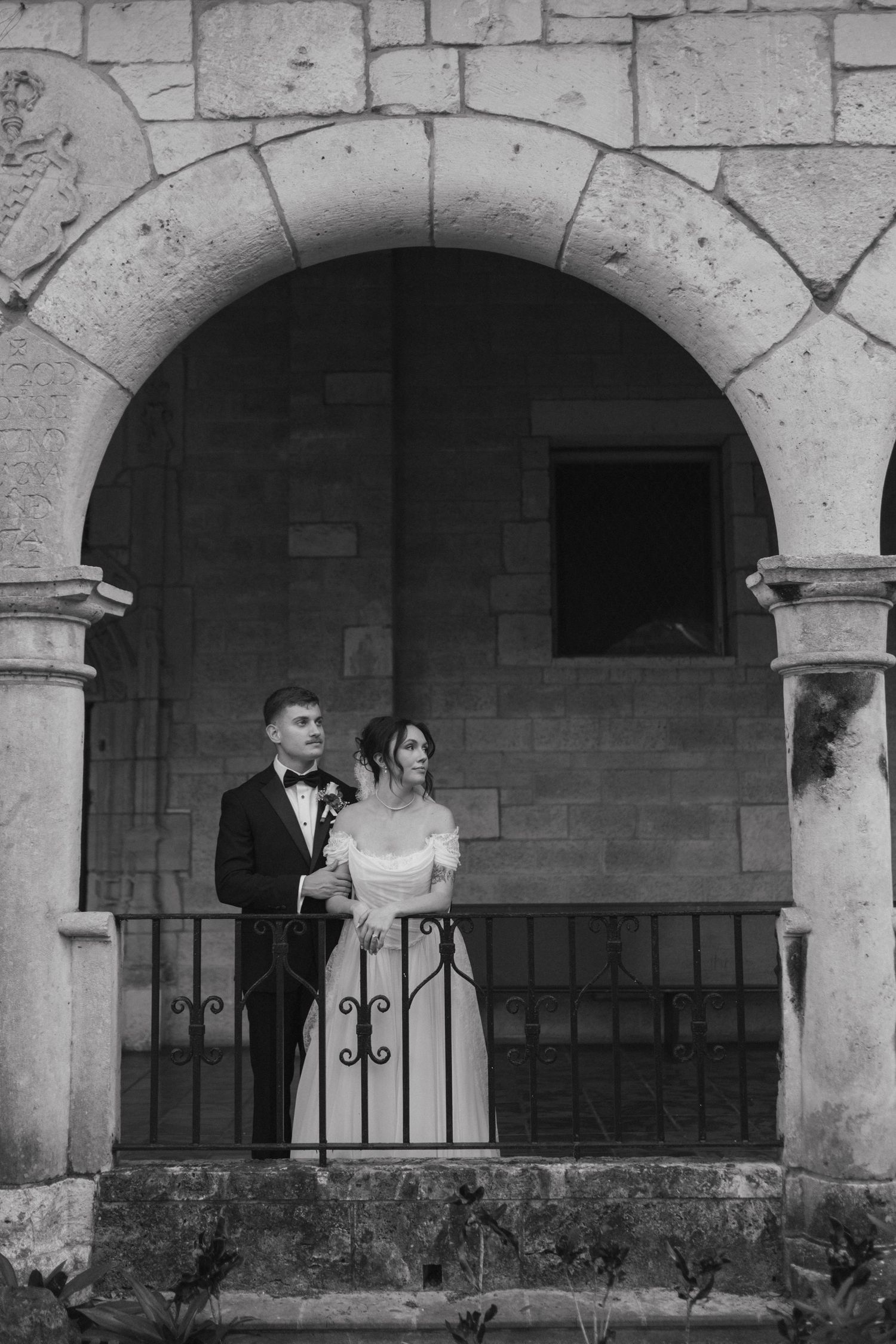 Black and white photo of elegant bride and groom standing under a stone arch on a balcony.