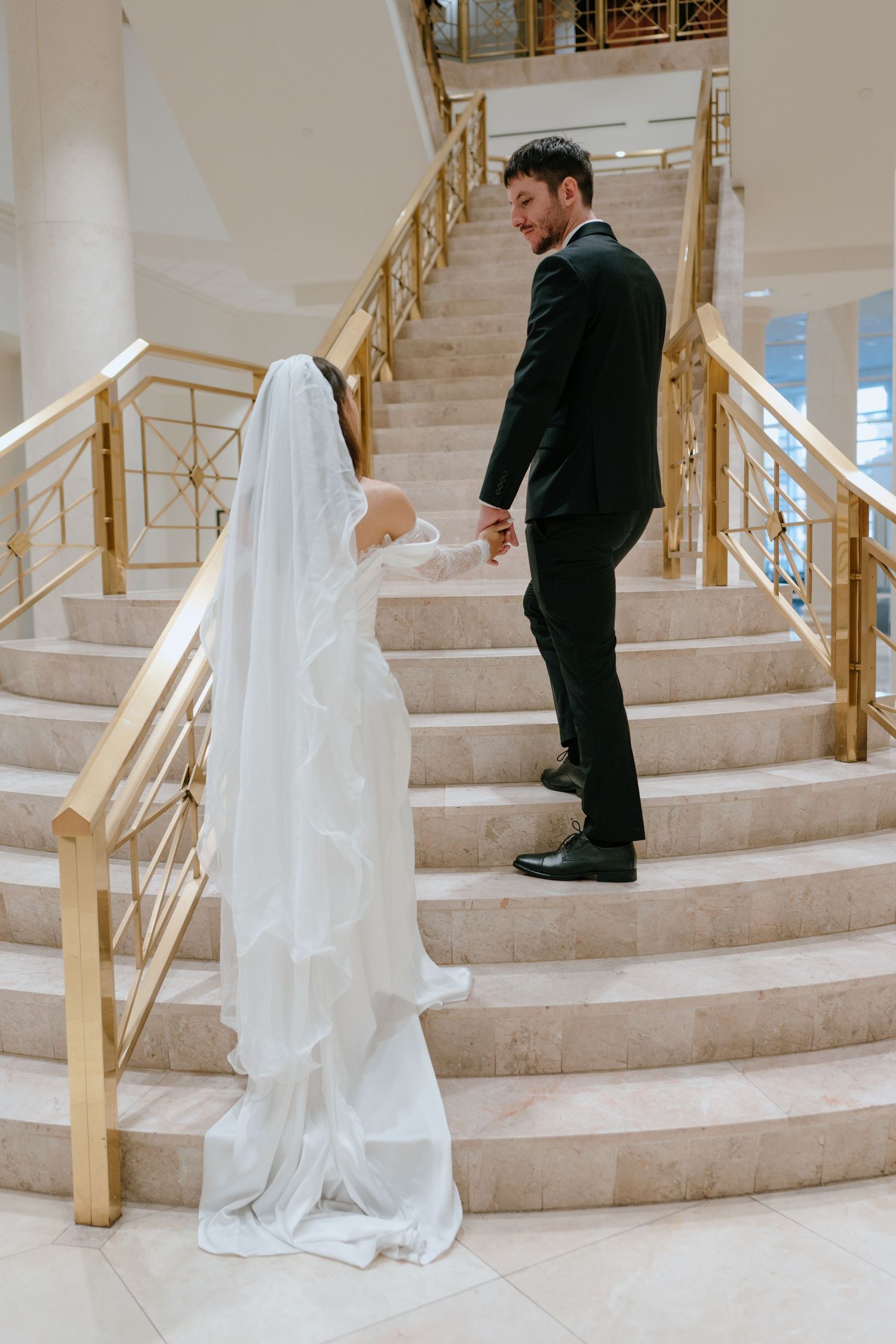 Bride and groom holding hands on grand marble staircase with gold railings.