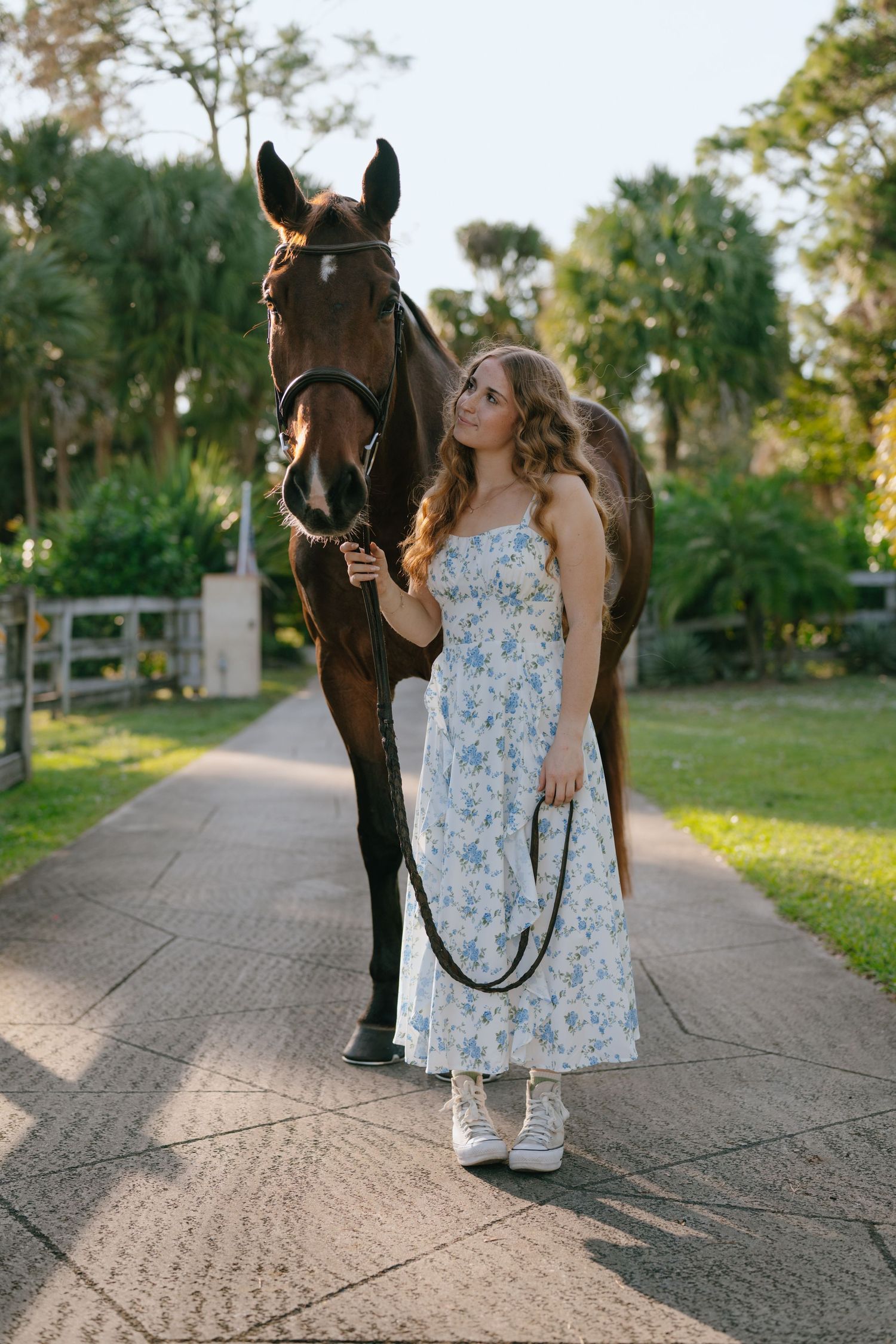 Young woman in floral sundress walks with dark bay horse on sunny stable pathway.