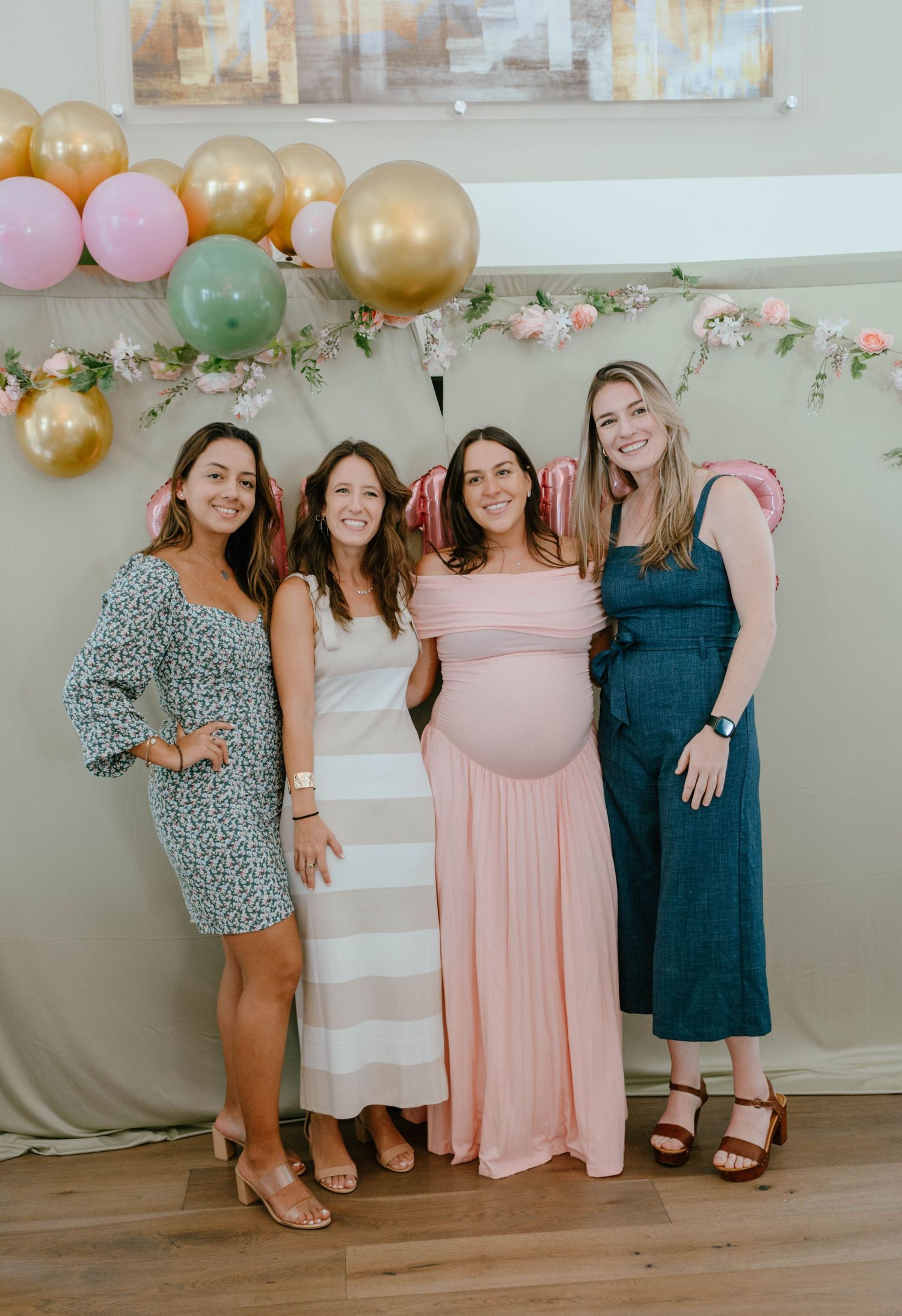Four women pose together at a baby shower, one visibly pregnant in a pink dress with balloon backdrop.