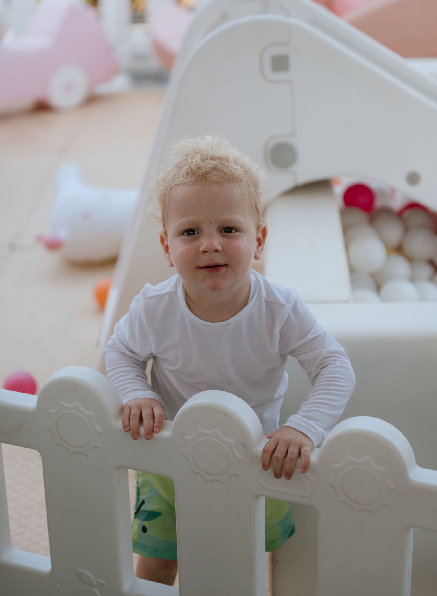 Curly-haired toddler in white shirt standing inside a white play fence at a children's birthday party.