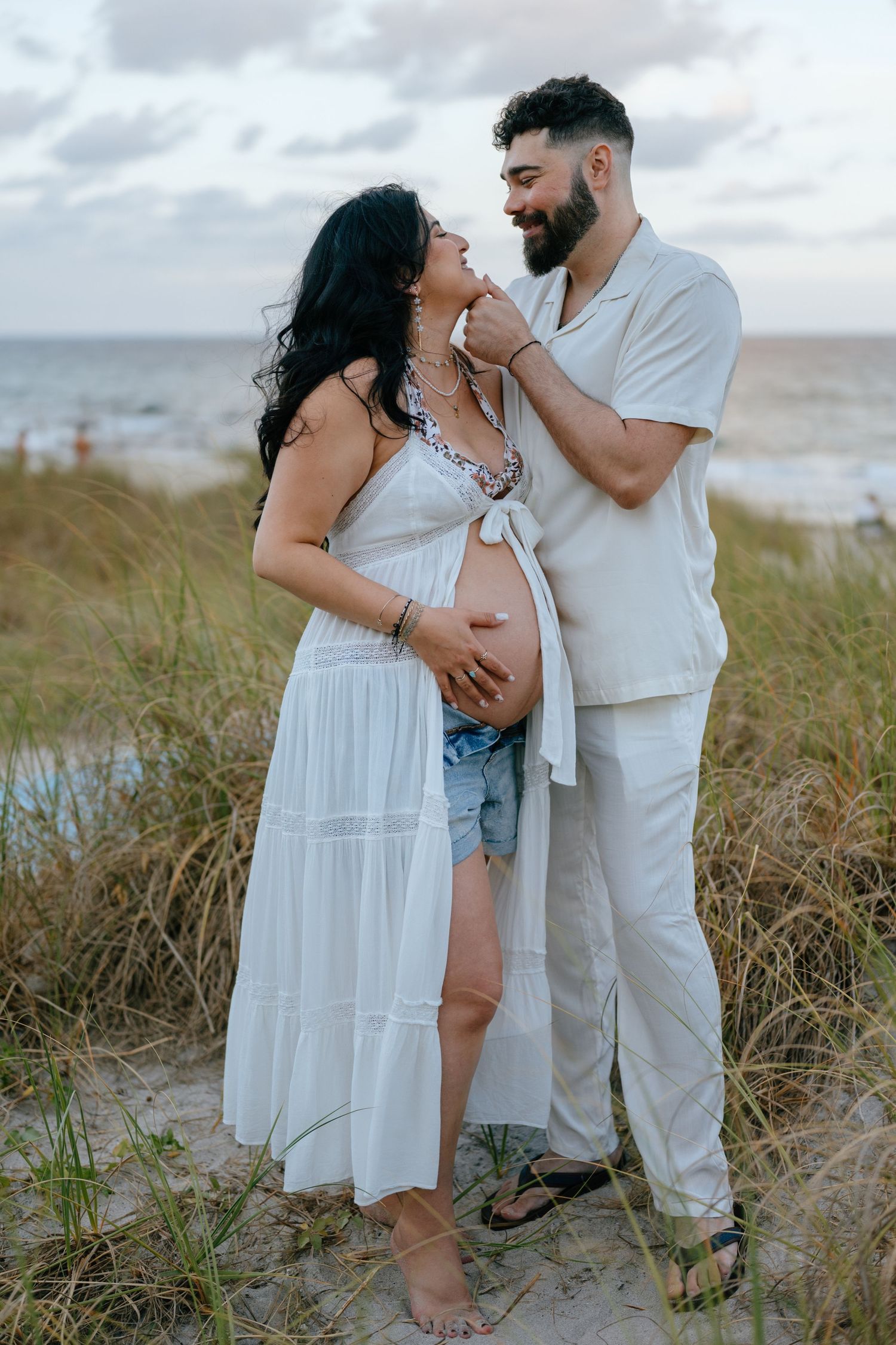 Couple sharing romantic moment on beach at sunset, pregnant woman in white dress.