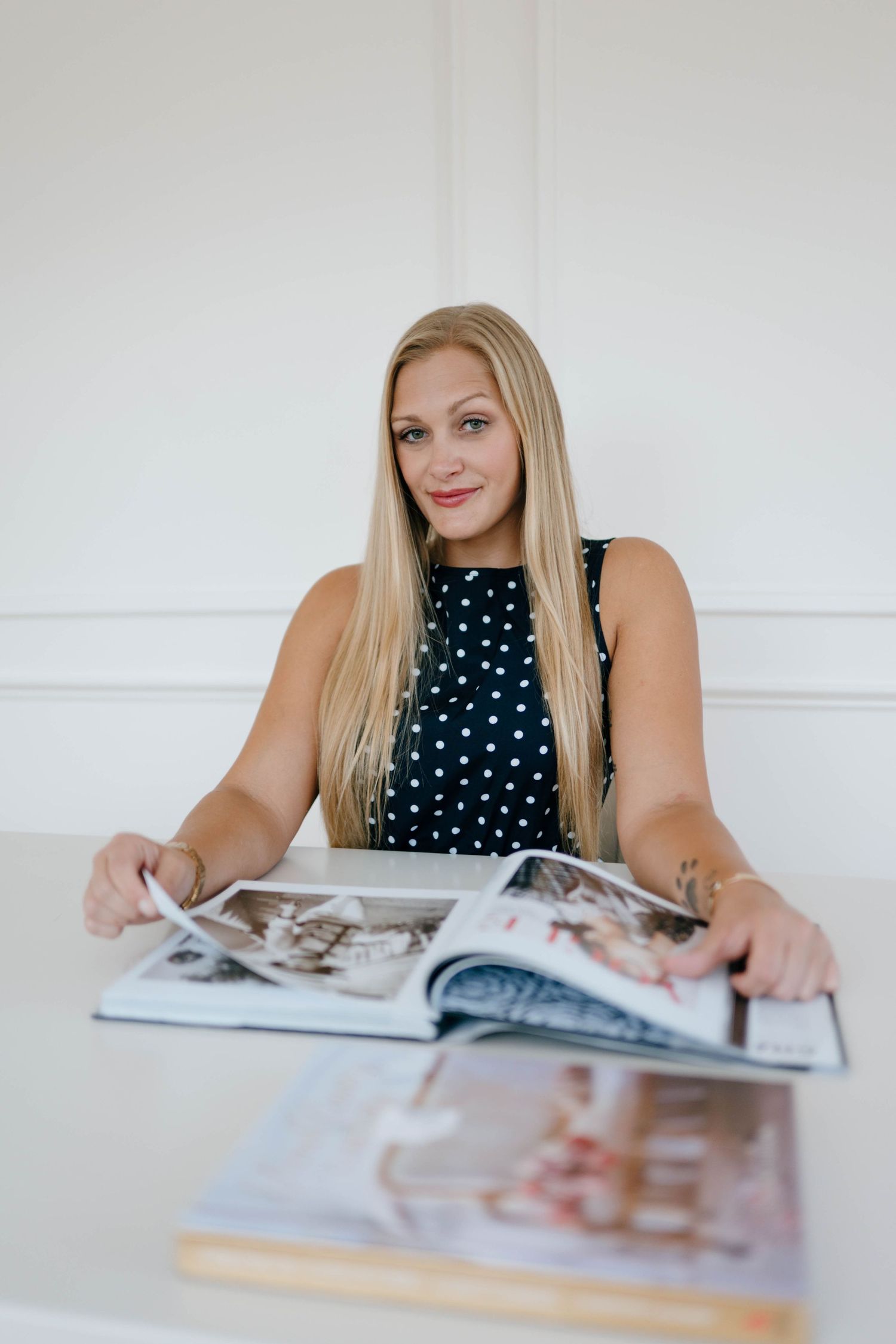 Smiling blonde woman in polka dot dress sitting at a white table reviewing large photo albums.