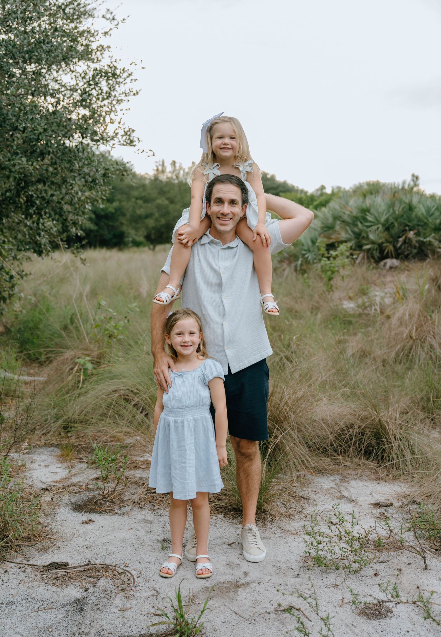 A father smiles with two young daughters, one on his shoulders, on a sandy path surrounded by tall grass.