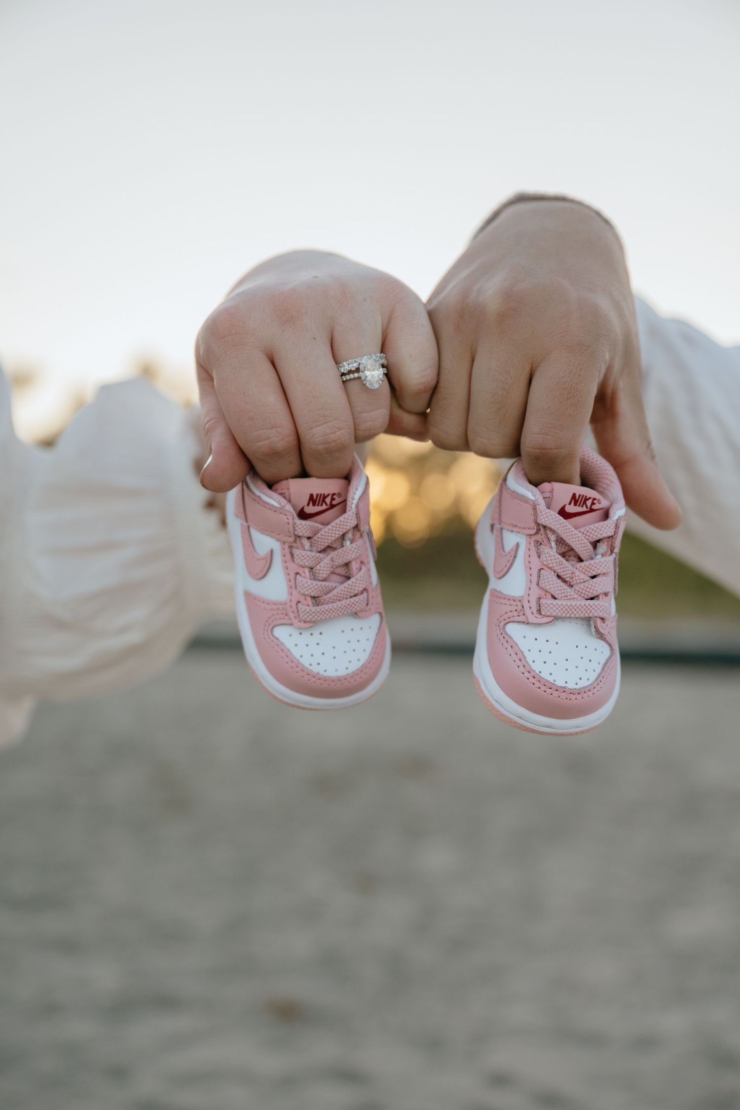 Hands holding tiny pink and white Nike baby sneakers, woman wearing diamond engagement ring.