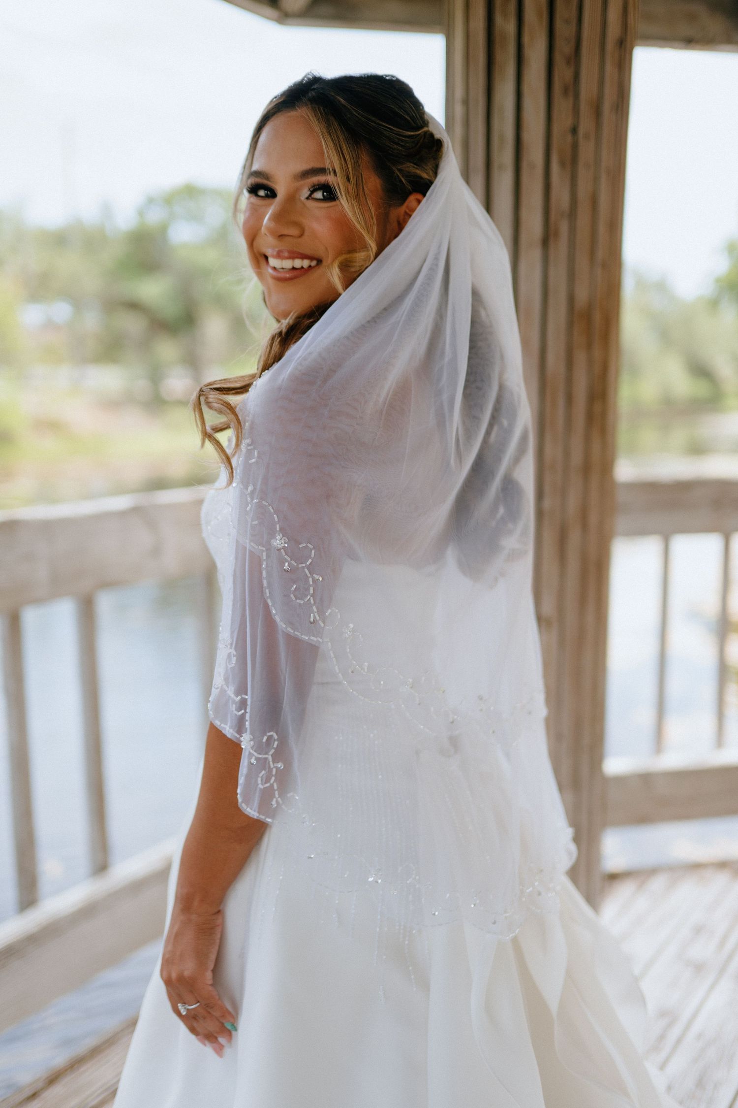 Smiling bride in a white gown with embroidered veil poses on a wooden deck outdoors.