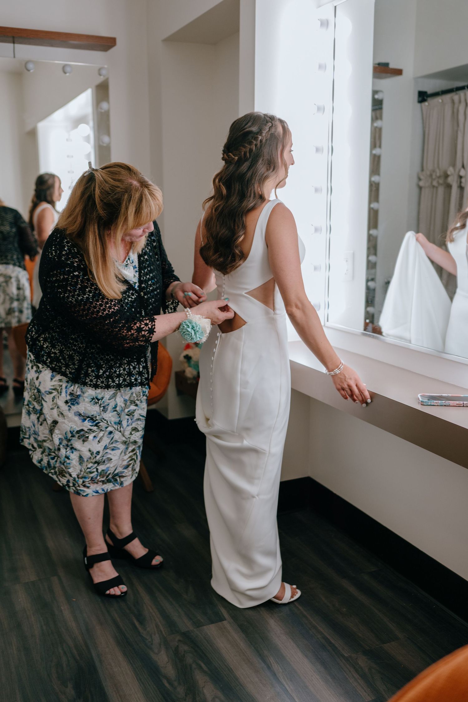 Mother helps bride fasten her elegant backless white wedding gown in a bridal suite before ceremony.