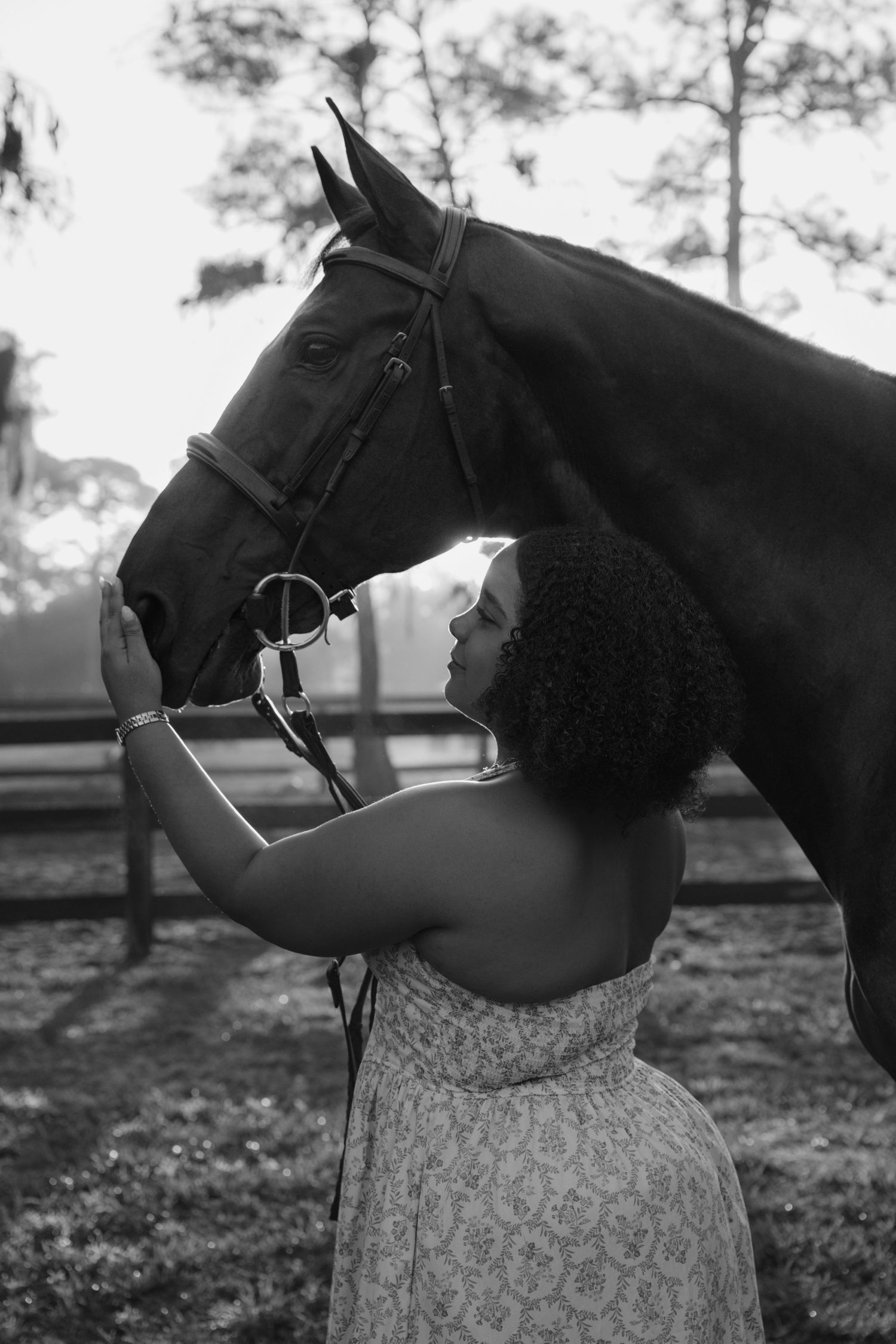 Black and white photo of a woman in a floral dress tenderly touching a dark horse's face.