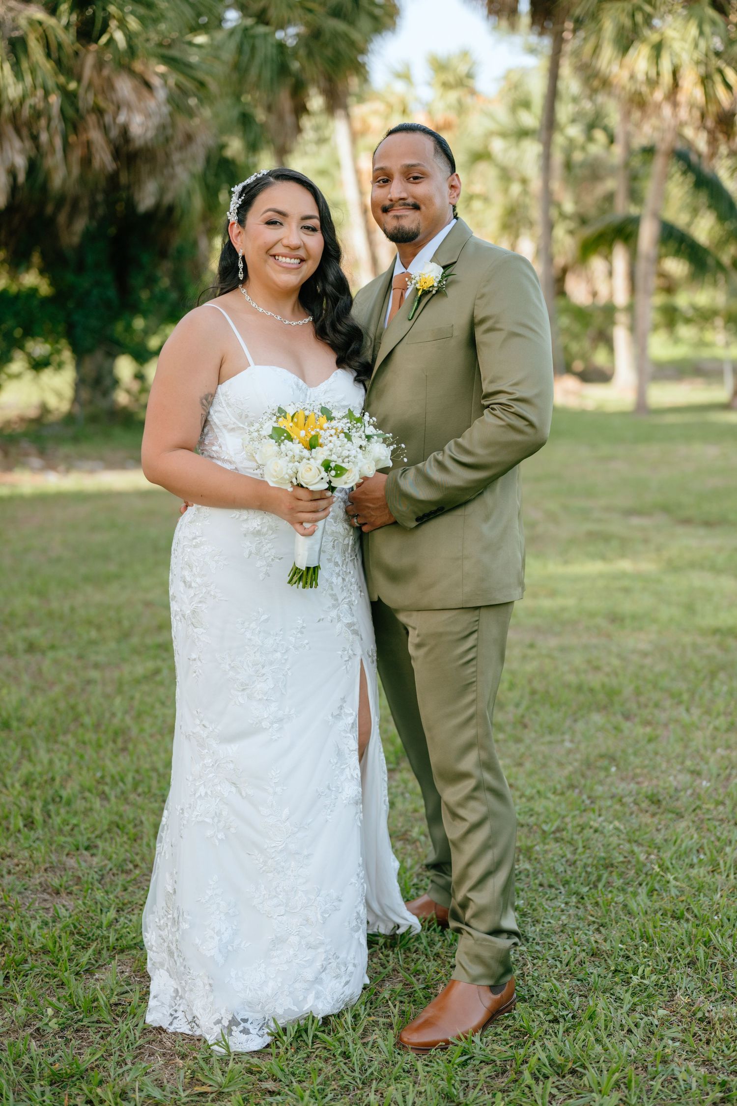 Bride and groom pose together outdoors among palm trees, she holds white bouquet in lace gown.