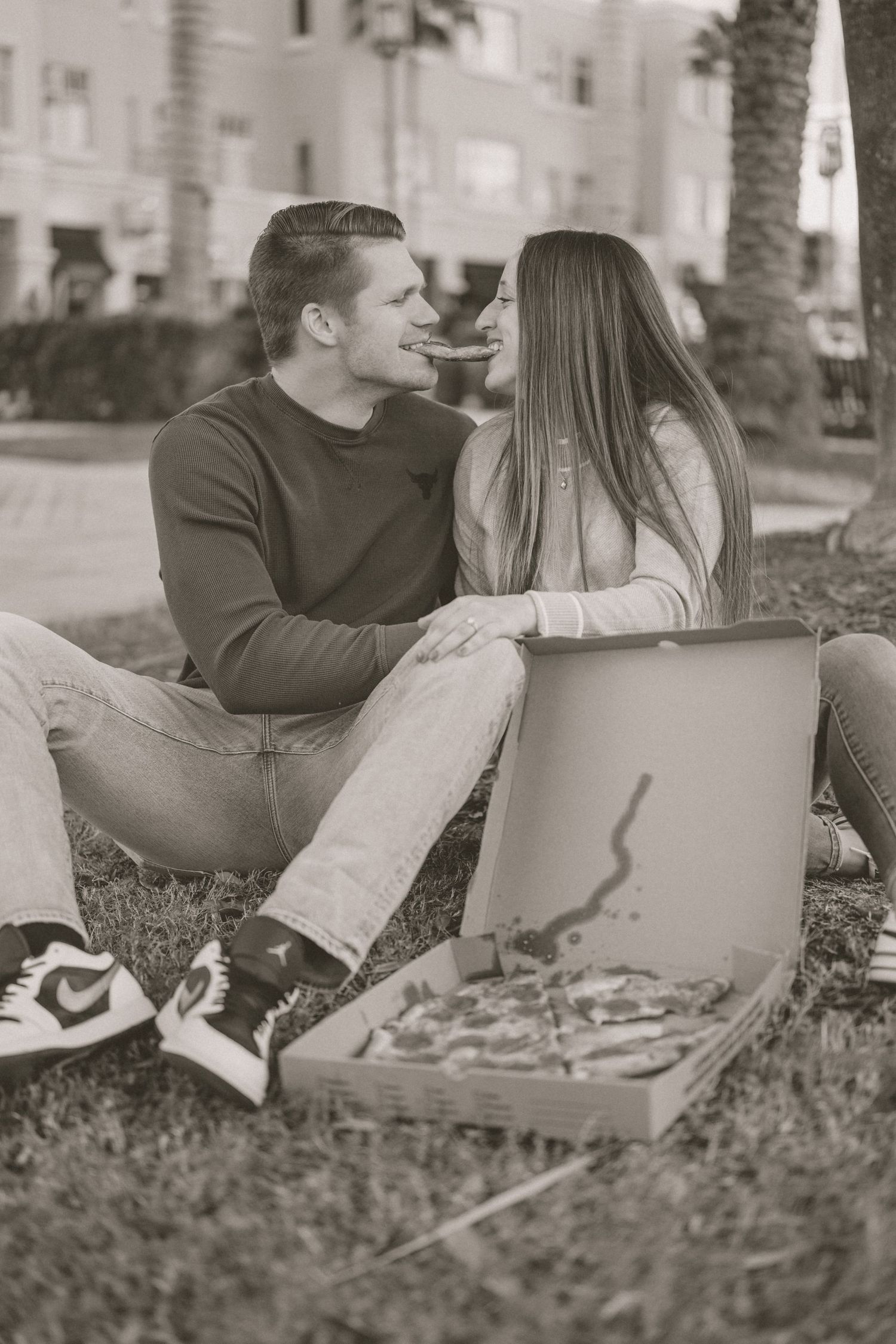 Romantic black and white photo of couple sharing pizza on grass in urban park setting.
