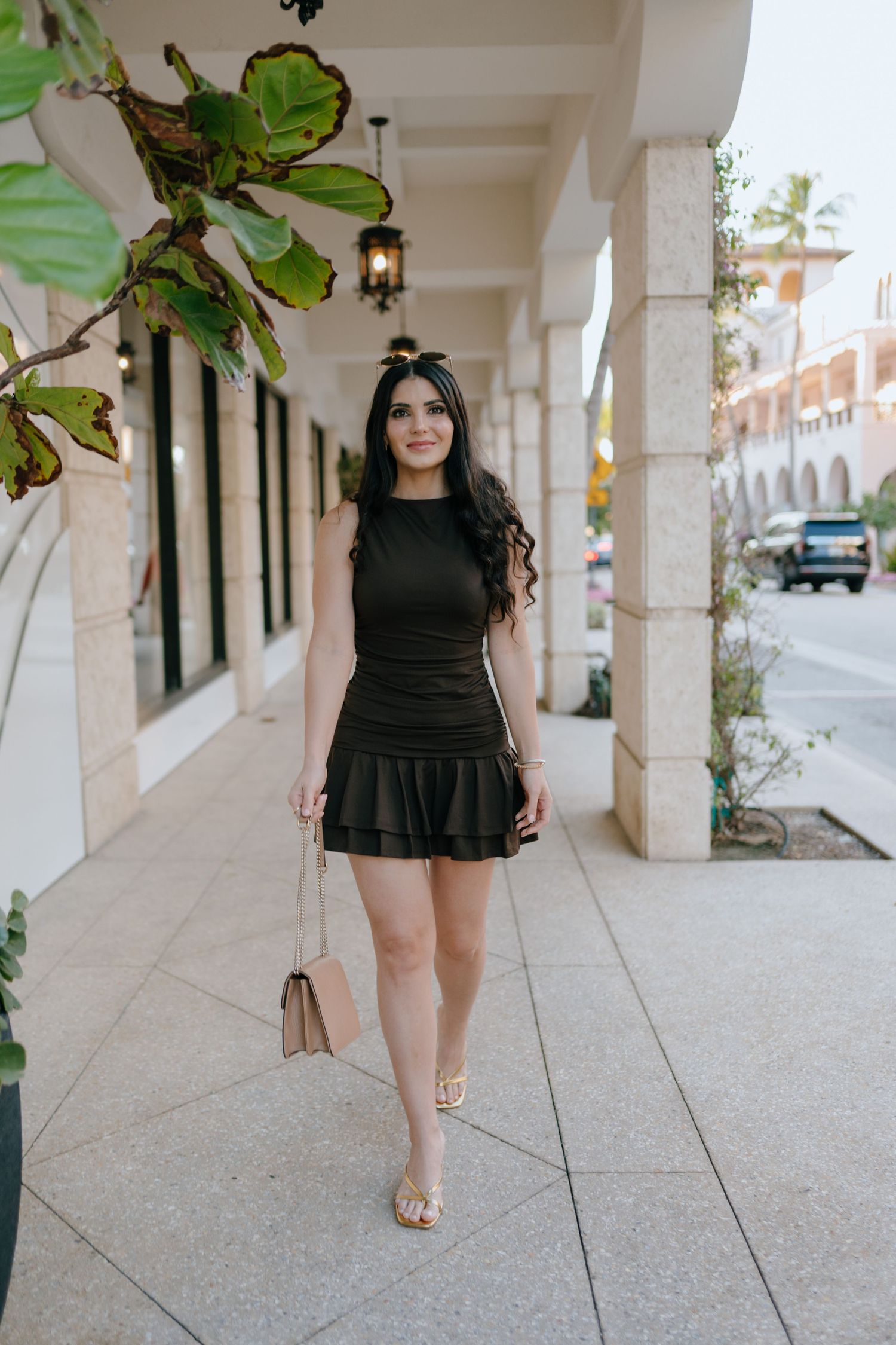 Woman in black ruched mini dress with ruffle hem walking along upscale outdoor shopping corridor.