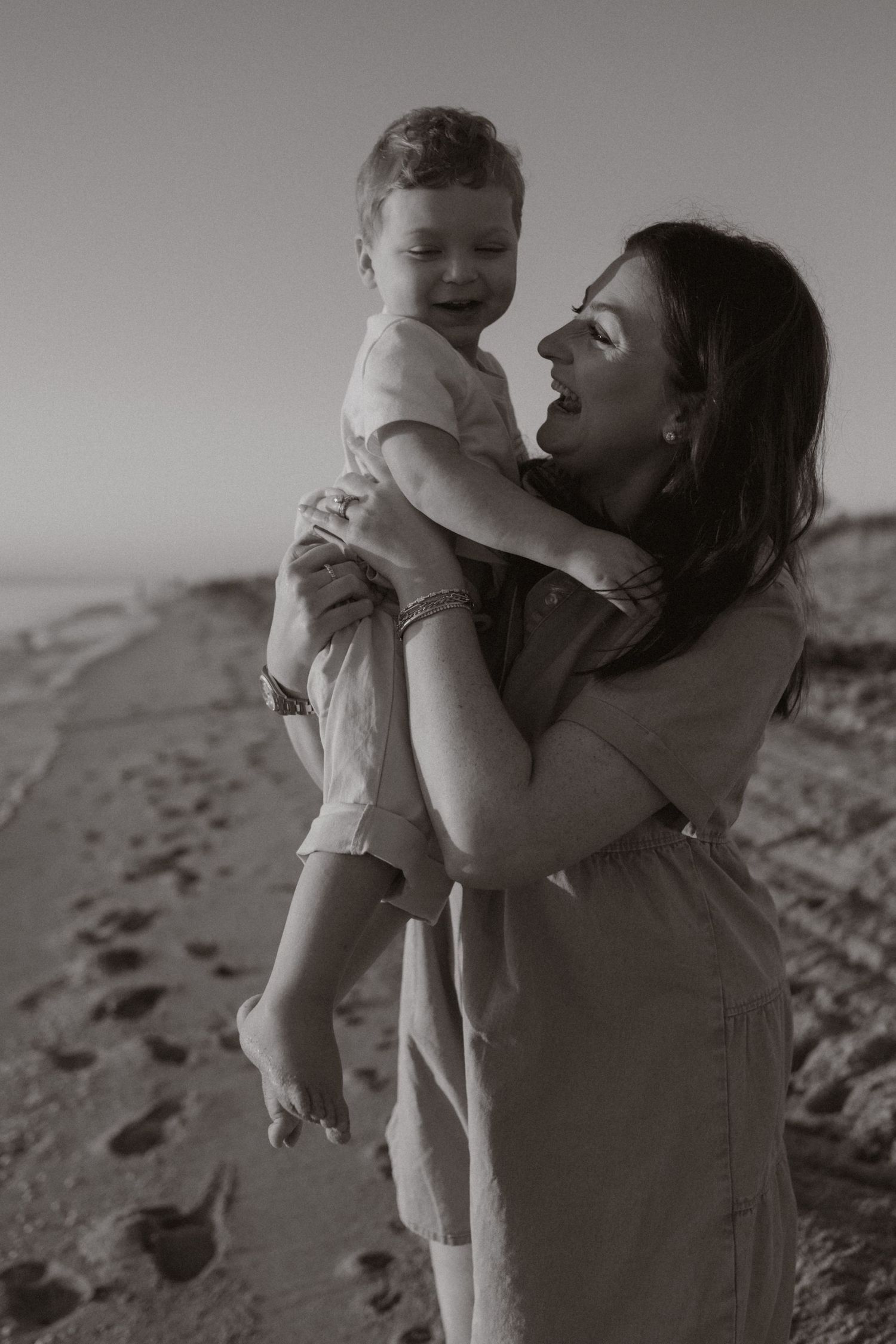 Black and white photo of a mother lifting her laughing baby on a sandy beach.