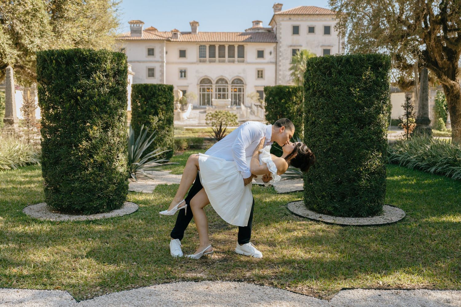 Couple shares a romantic dip kiss surrounded by manicured hedges outside a grand villa.