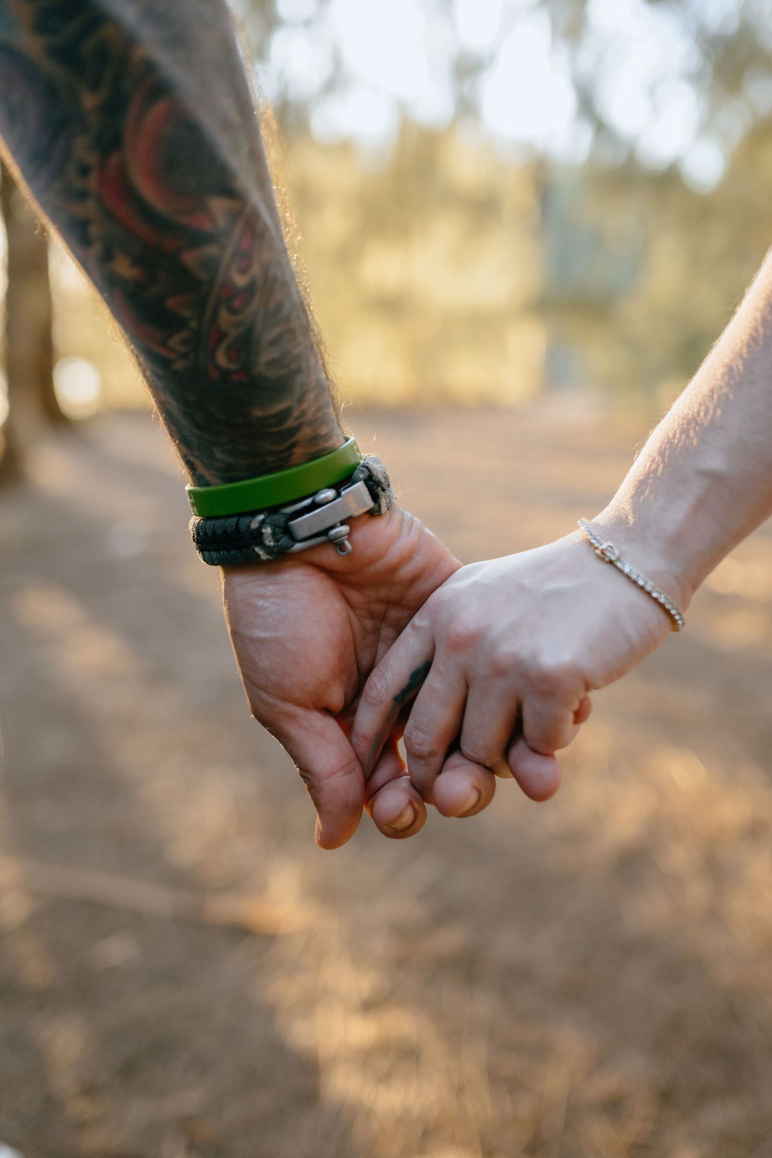 Close-up of couple holding hands, man with tattoed sleeve and green bracelet, woman with delicate chain bracelet.