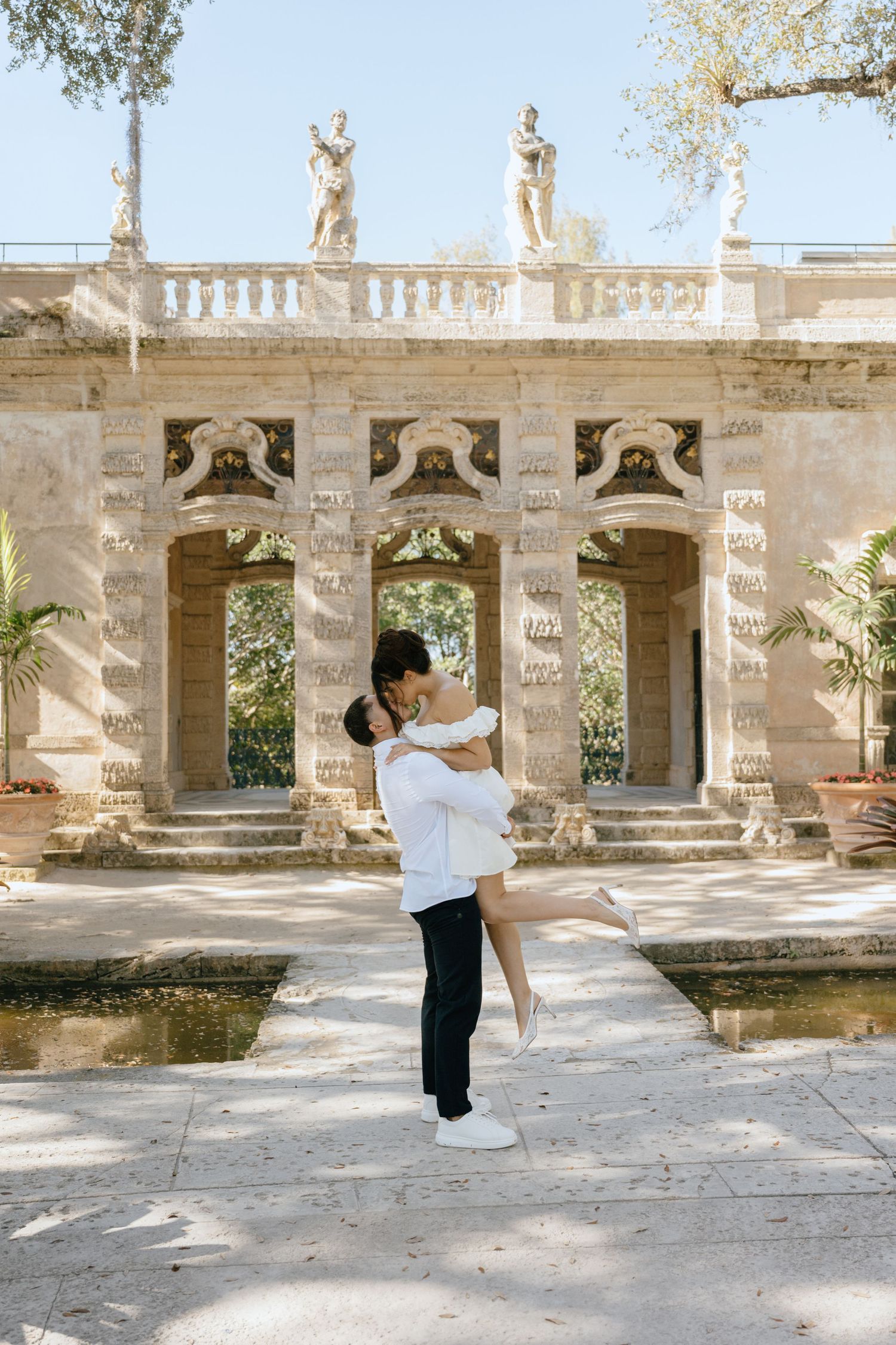 Man lifting woman in white dress in front of ornate stone arches and reflecting pool at historic estate.