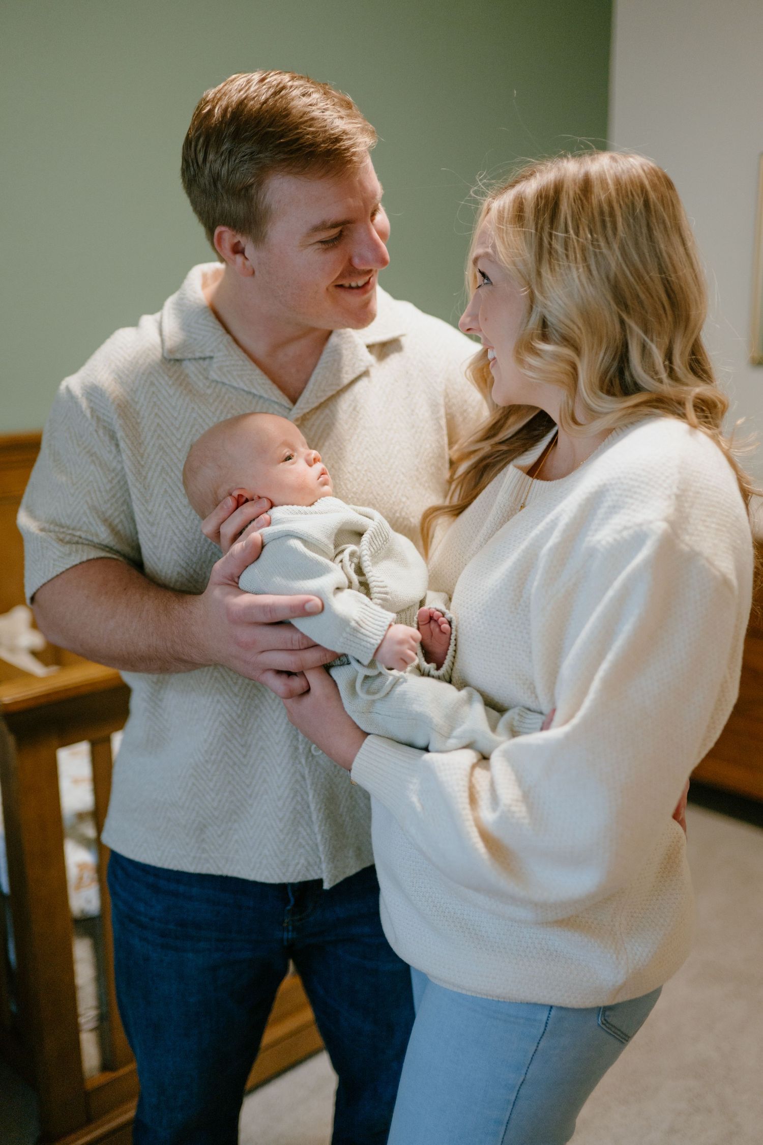 A smiling couple in casual outfits lovingly holding their newborn baby together indoors.