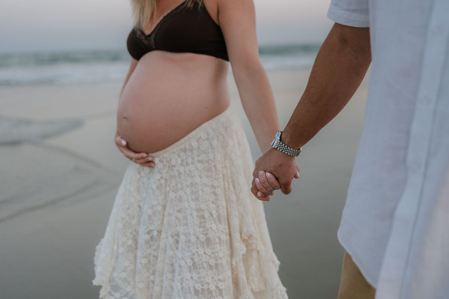Pregnant woman in white lace skirt holding partner's hand on ocean beach shoreline.