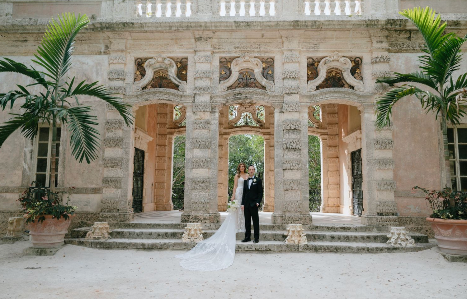 Bride and groom standing beneath ornate stone arches of a grand historic villa wedding venue with palm trees.