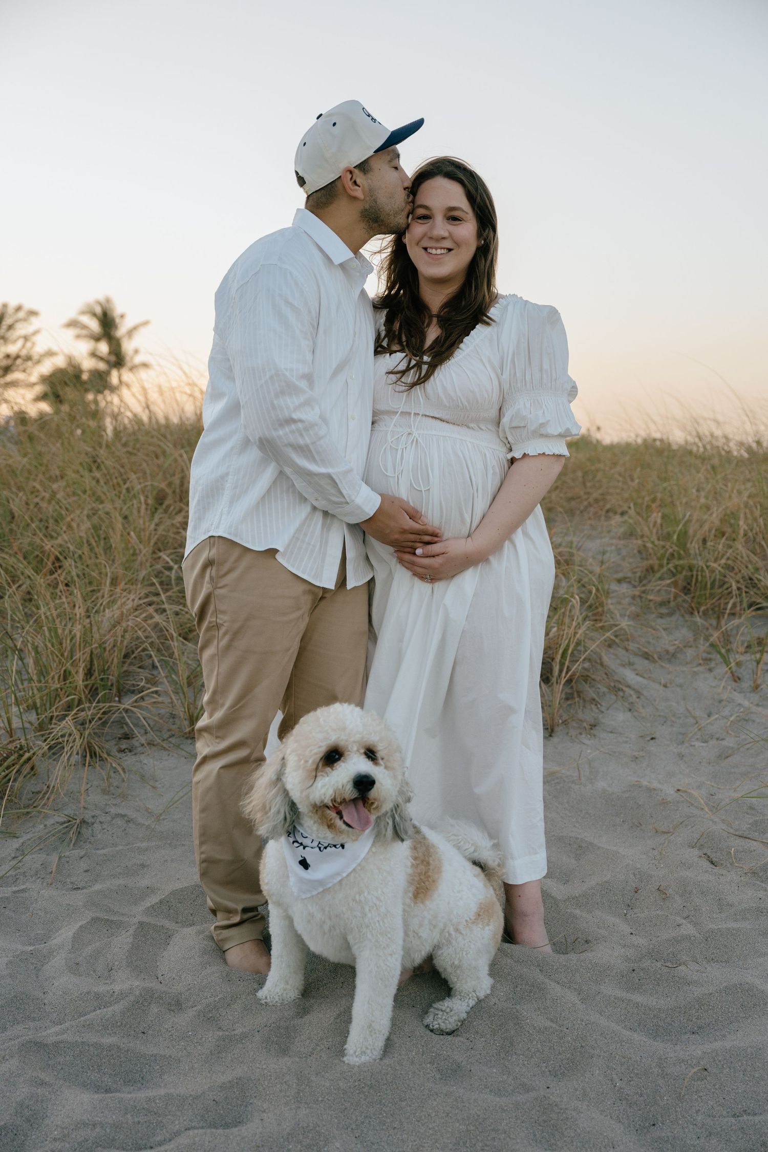 Man kissing pregnant woman's cheek on sandy beach dunes with white fluffy dog wearing bandana.
