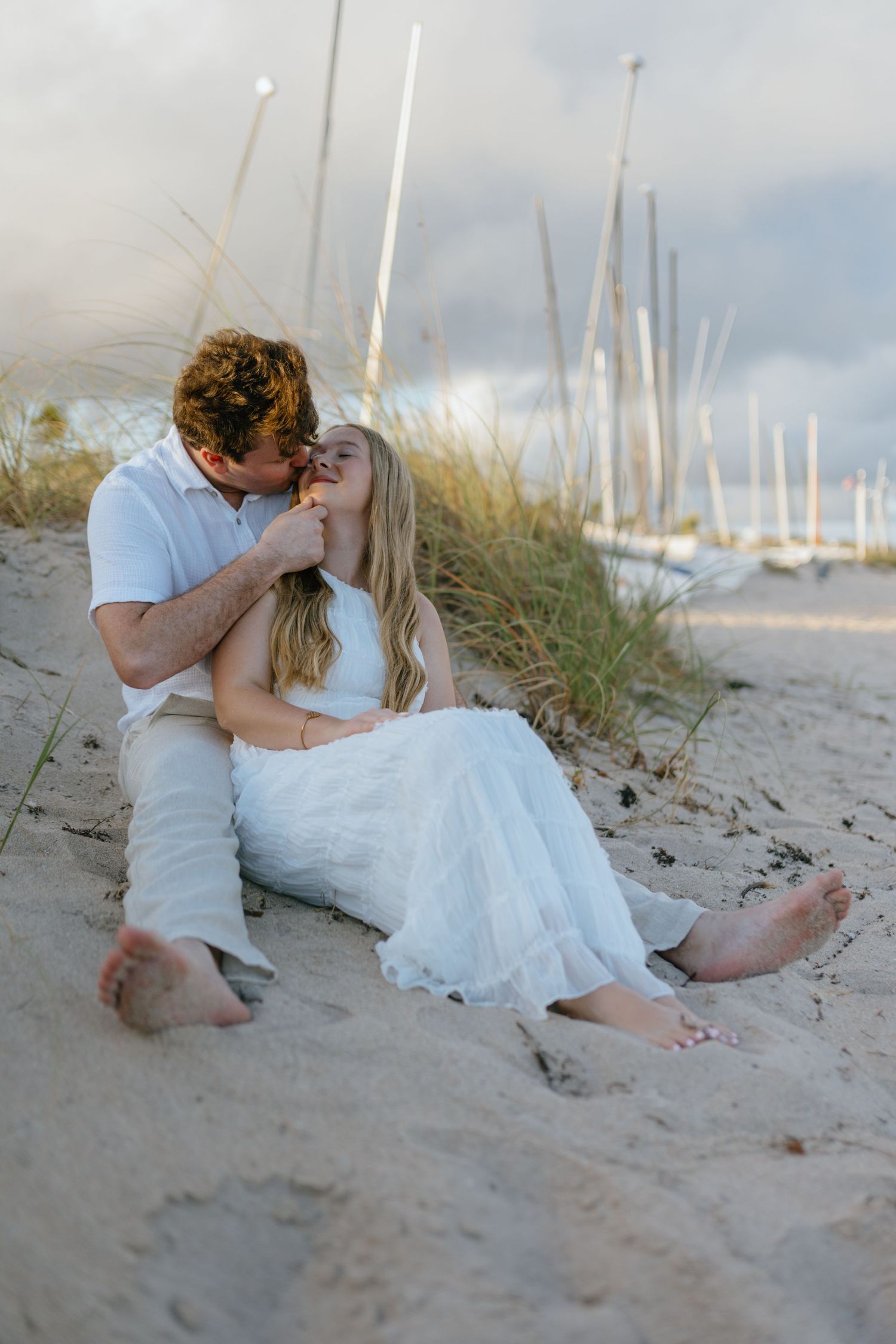Couple sitting barefoot on sandy beach dunes at sunset, sharing a romantic kiss during engagement photos.