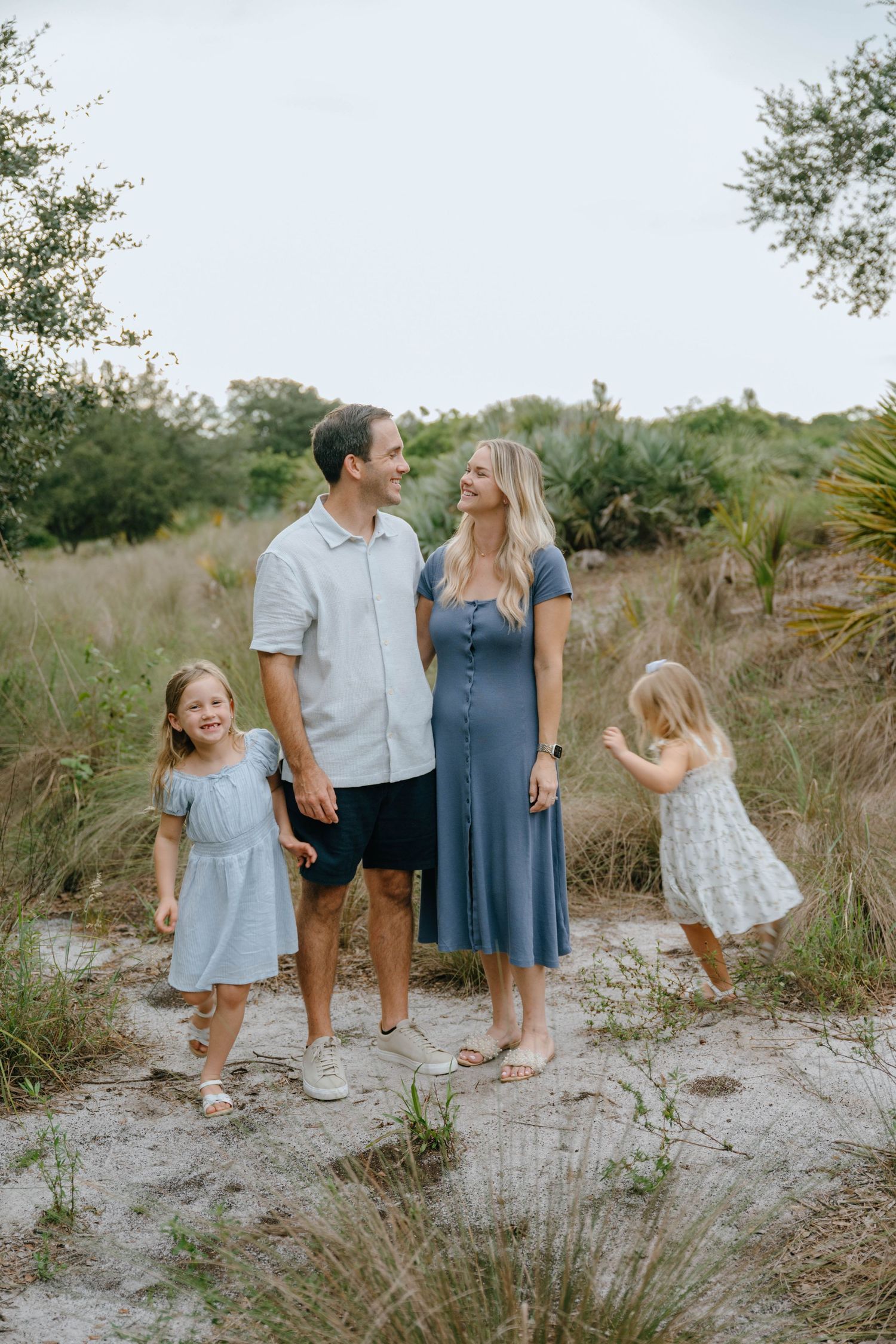 A happy family of four walks together on a sandy trail surrounded by lush green vegetation.