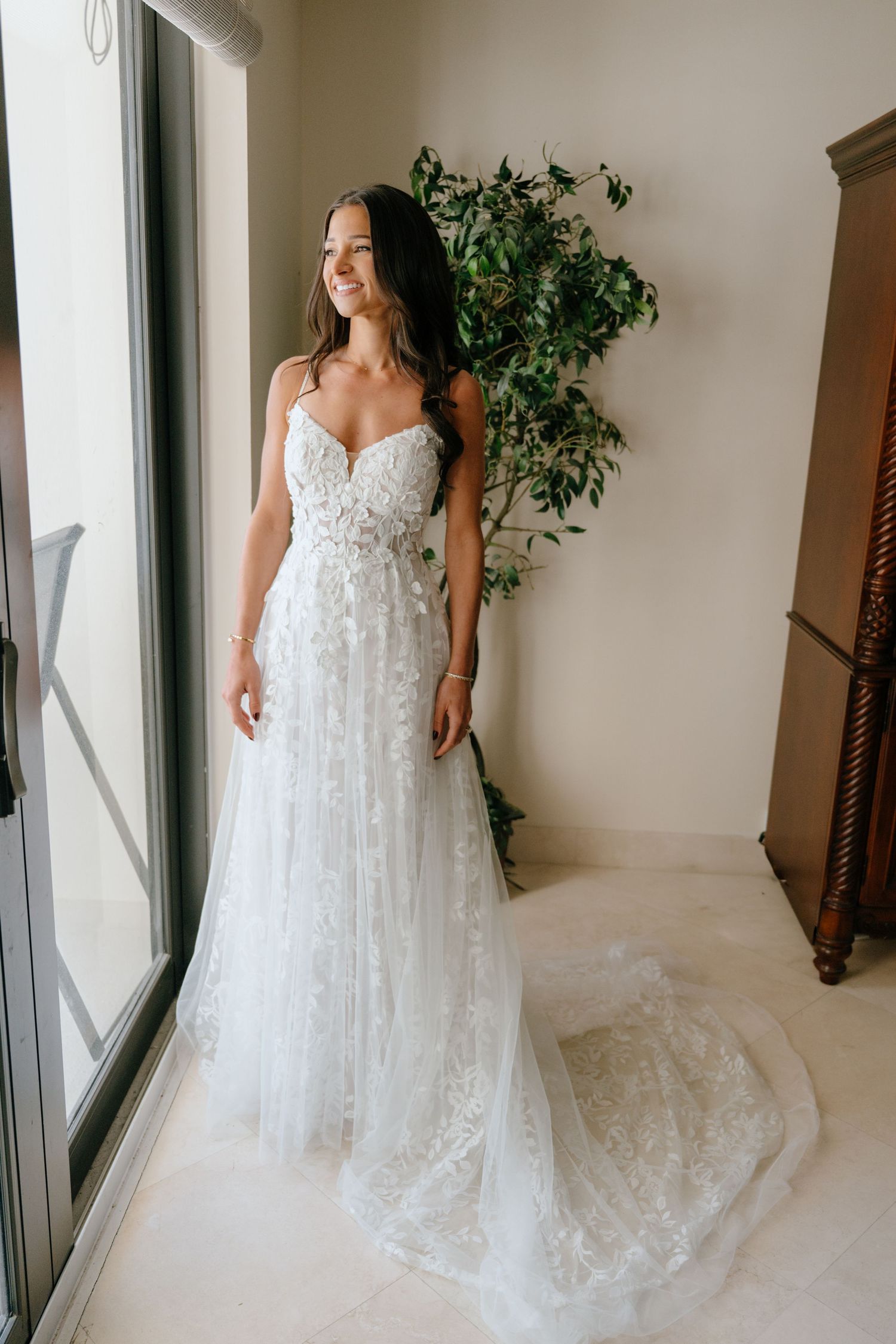 Smiling bride in a floral lace A-line wedding gown standing by a window with greenery backdrop.