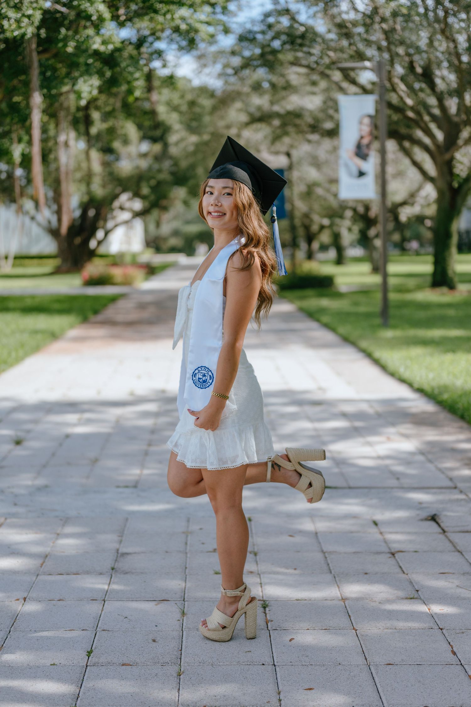 Joyful graduate posing in white mini dress, black cap, and platform heels on a tree-lined campus path.