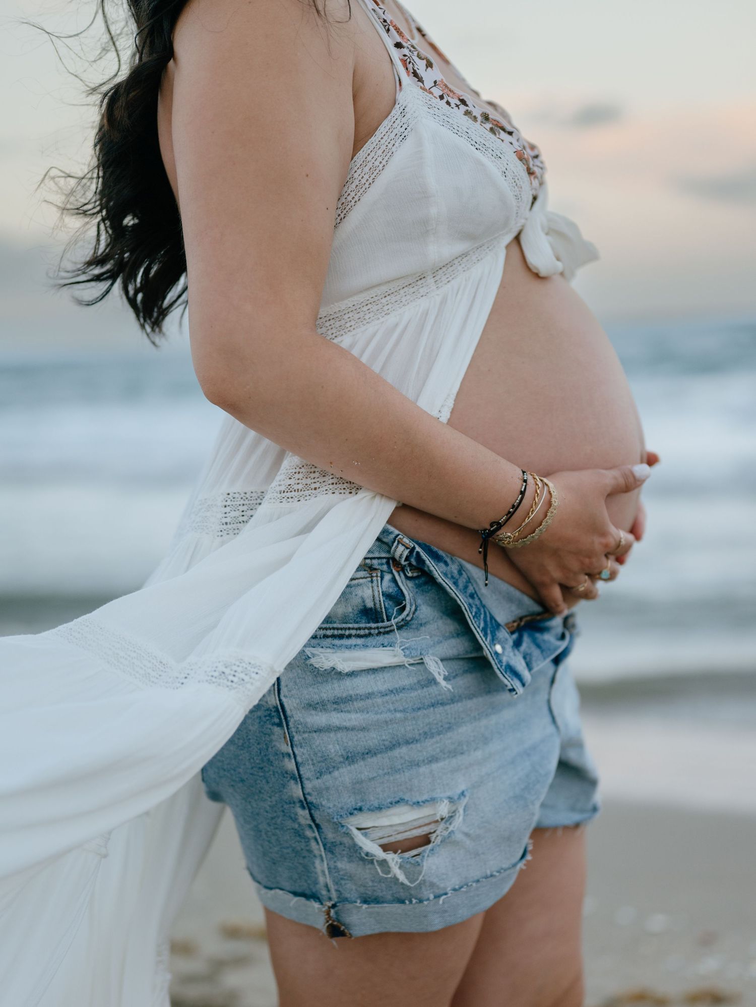 Pregnant woman in white top and denim shorts holding baby bump on beach at dusk.