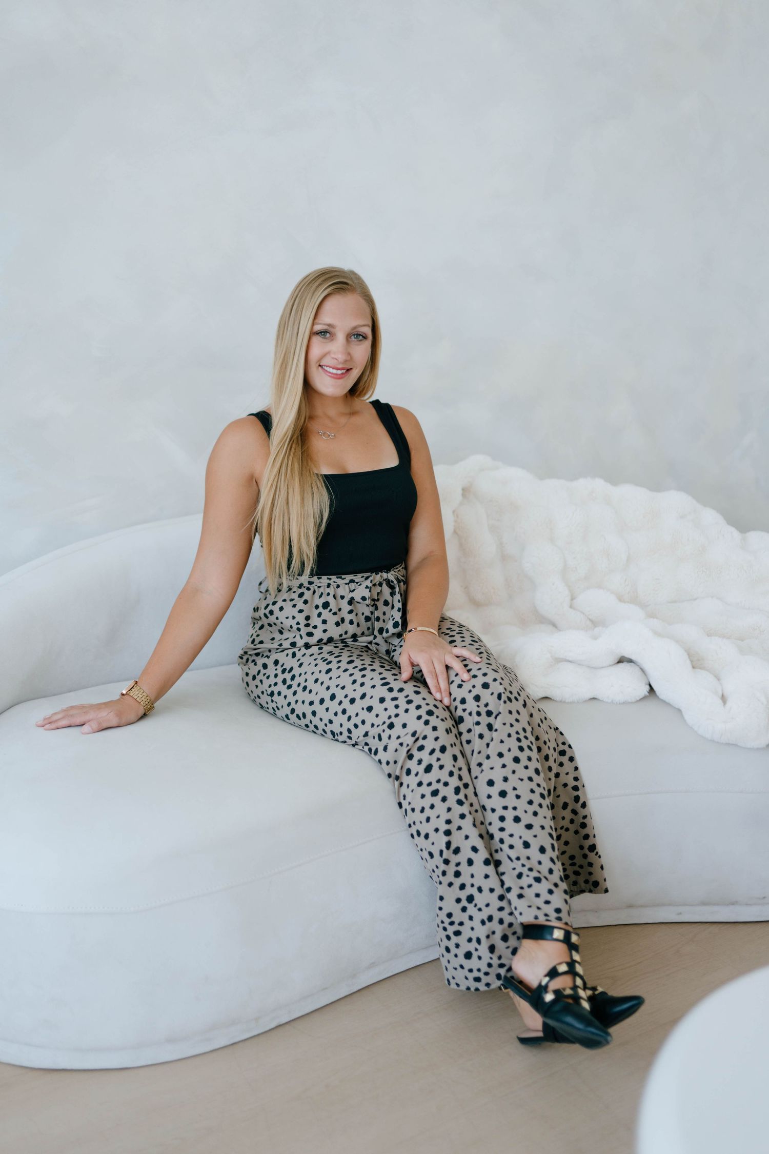 Blonde woman in black top and leopard print pants sitting on white curved sofa.