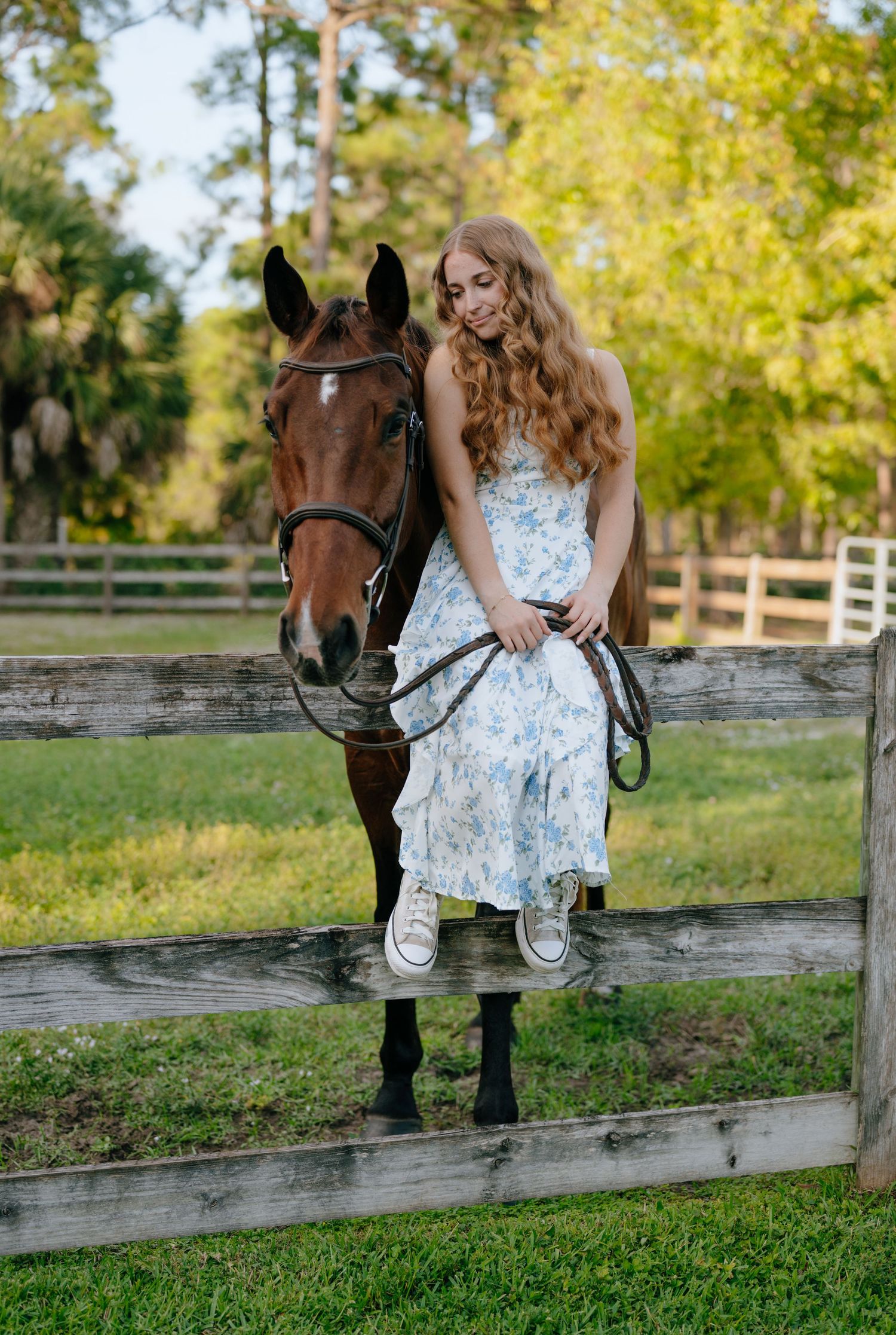 Young woman in floral dress sitting on wooden fence with brown horse at sunny equestrian farm.