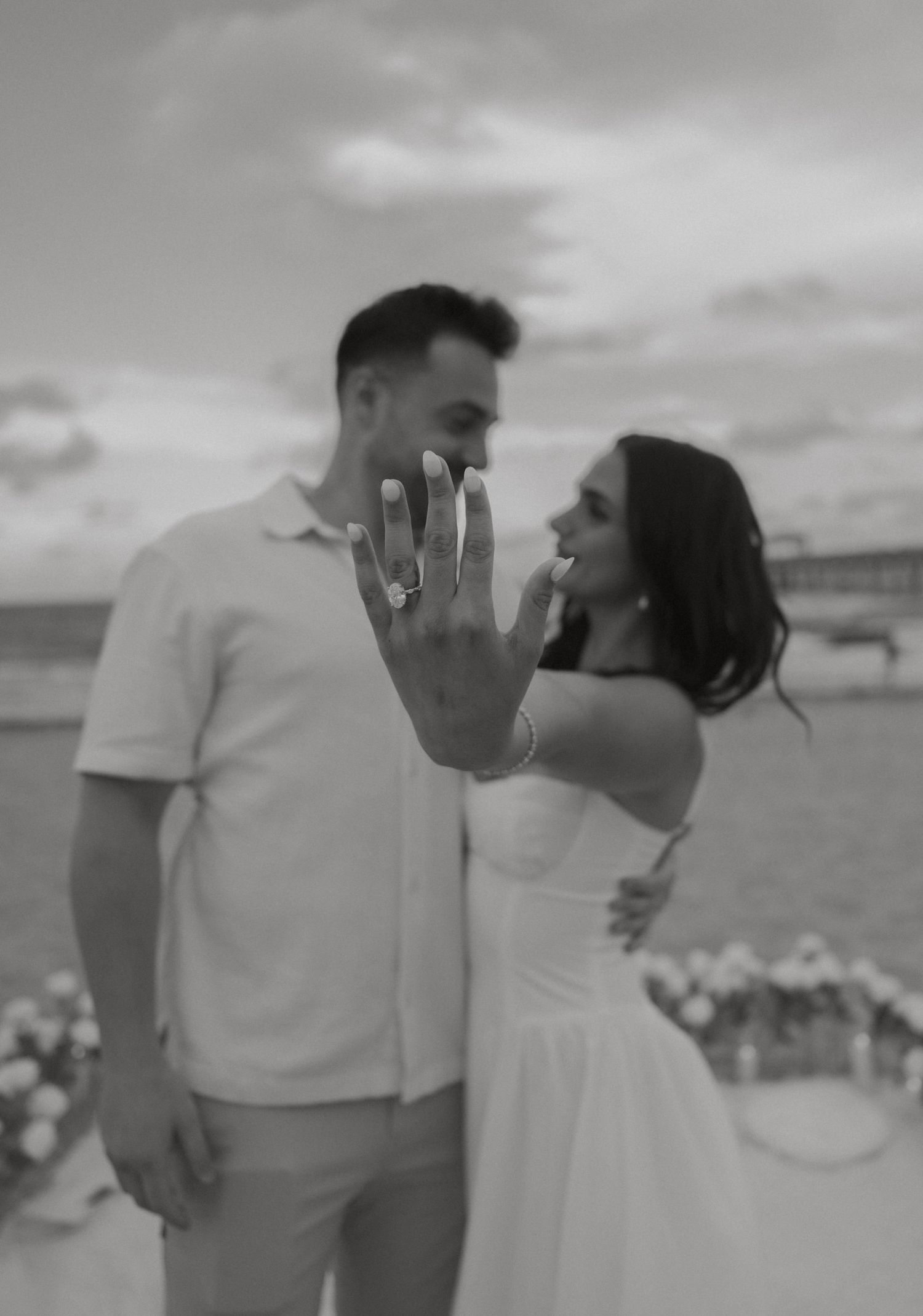 Black and white photo of smiling couple showing off engagement ring on beach after proposal.