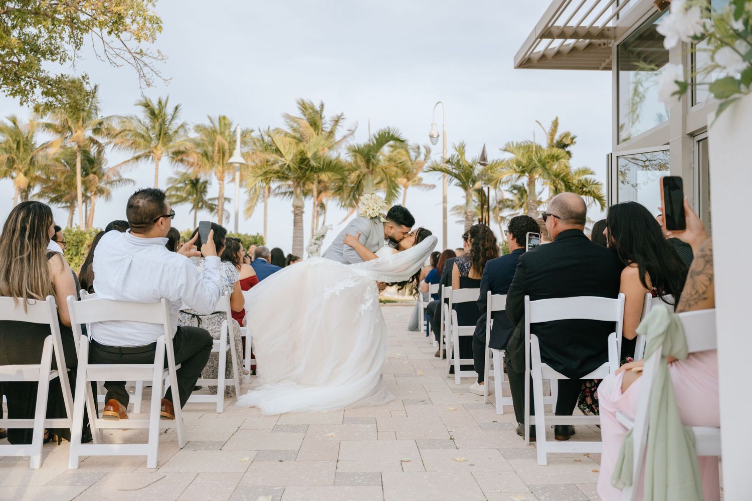 Outdoor beach wedding ceremony with bride and groom kissing at the altar amid palm trees.