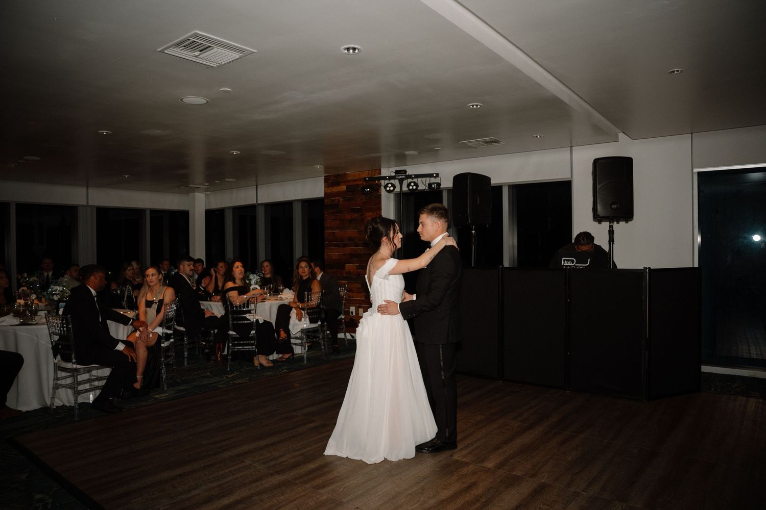 Bride and groom sharing their first dance before seated wedding guests in a dimly lit reception hall.