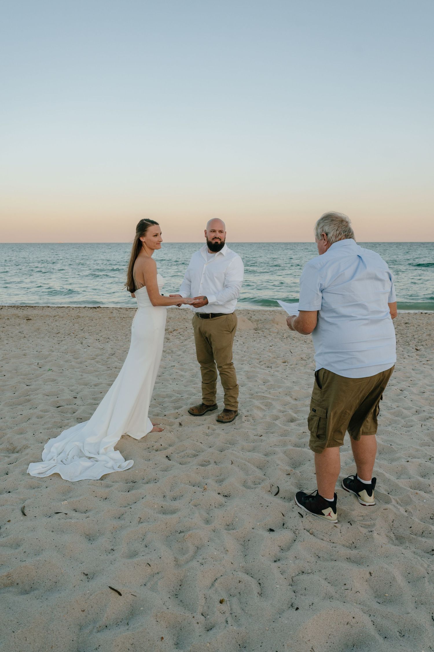 Bride and groom exchanging vows with officiant on a sandy beach at twilight.