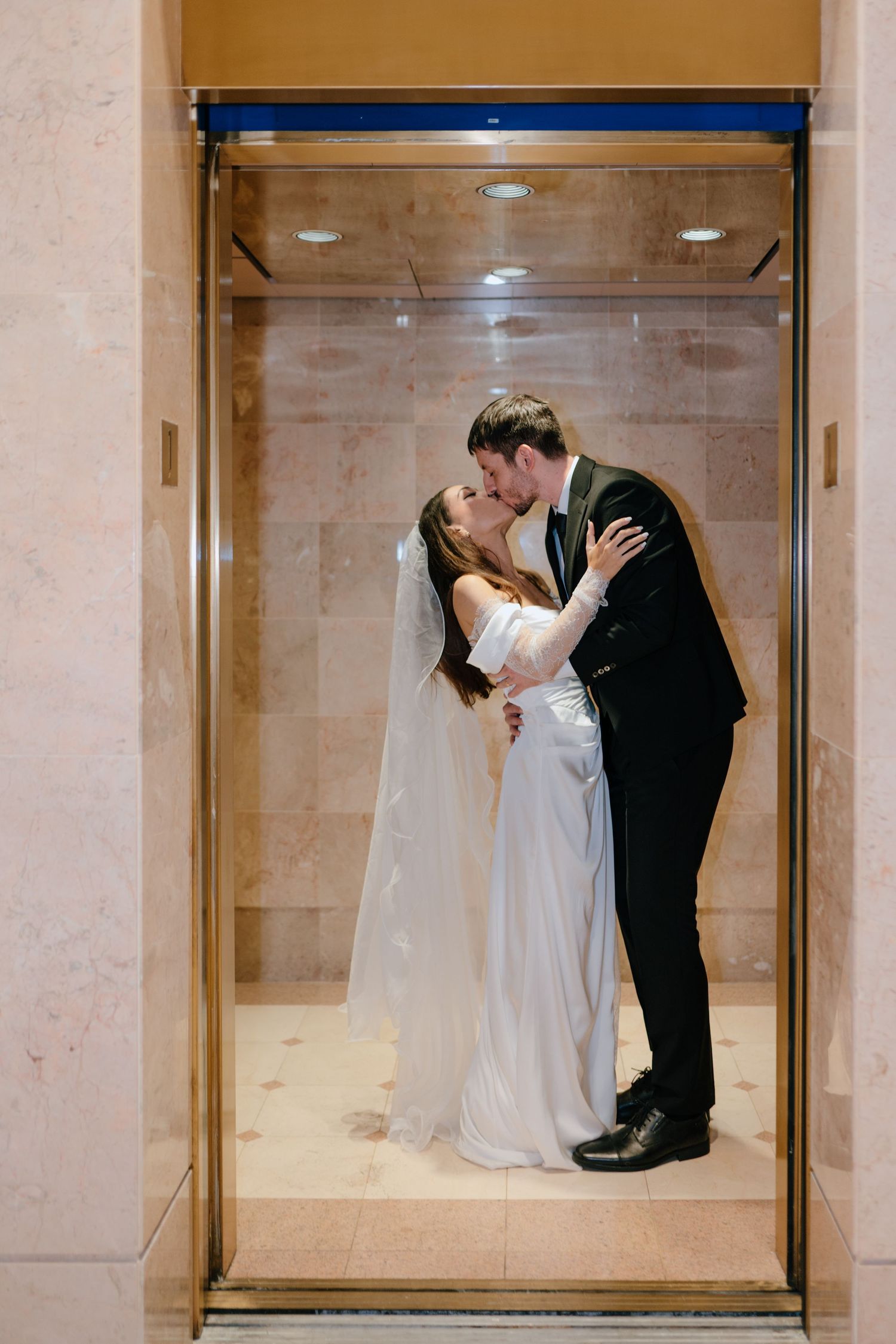 Bride and groom sharing a kiss inside a gold-framed elevator at wedding venue.