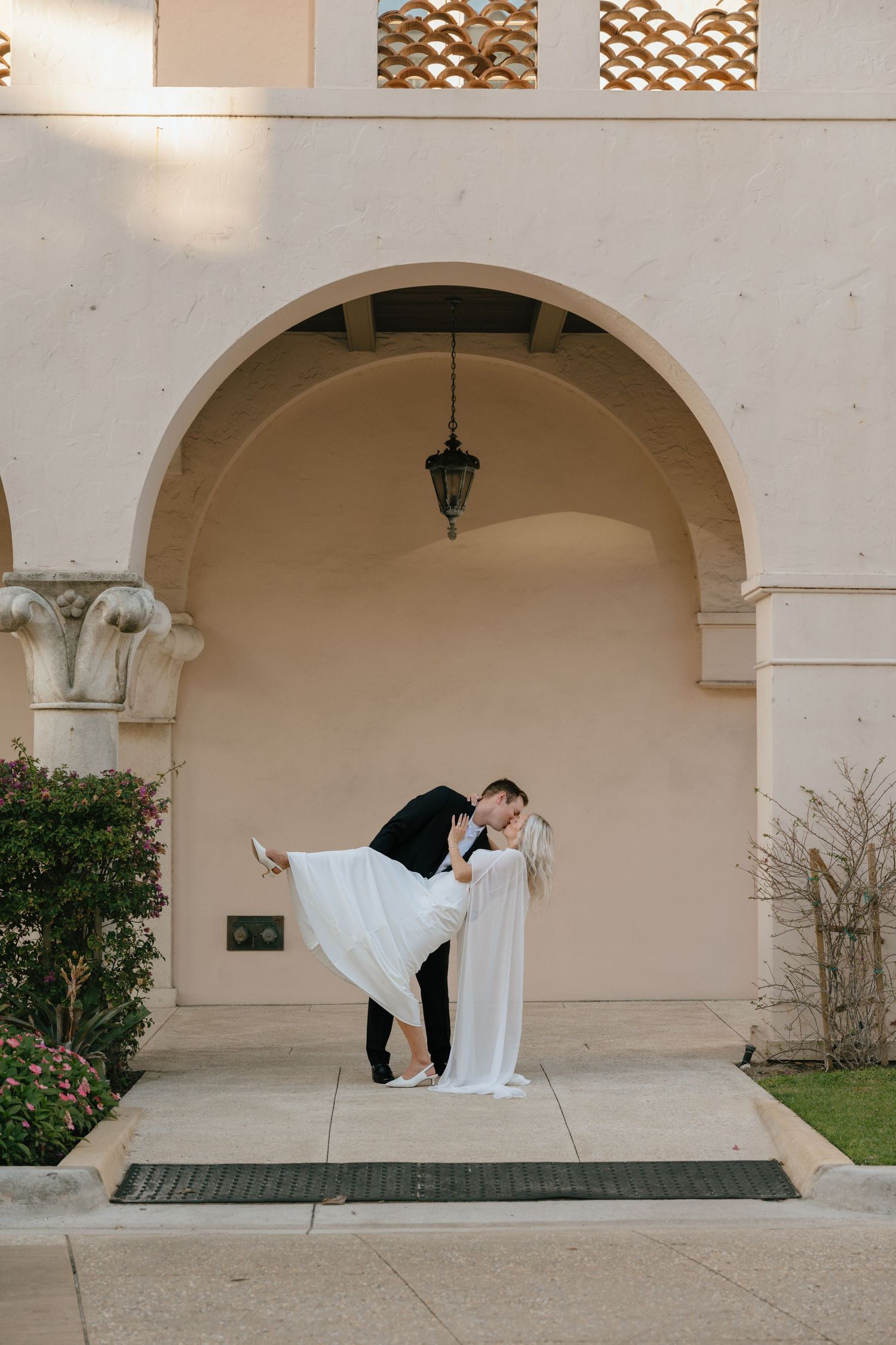 Groom dips bride in flowing white gown for a romantic kiss beneath an elegant stone arch.