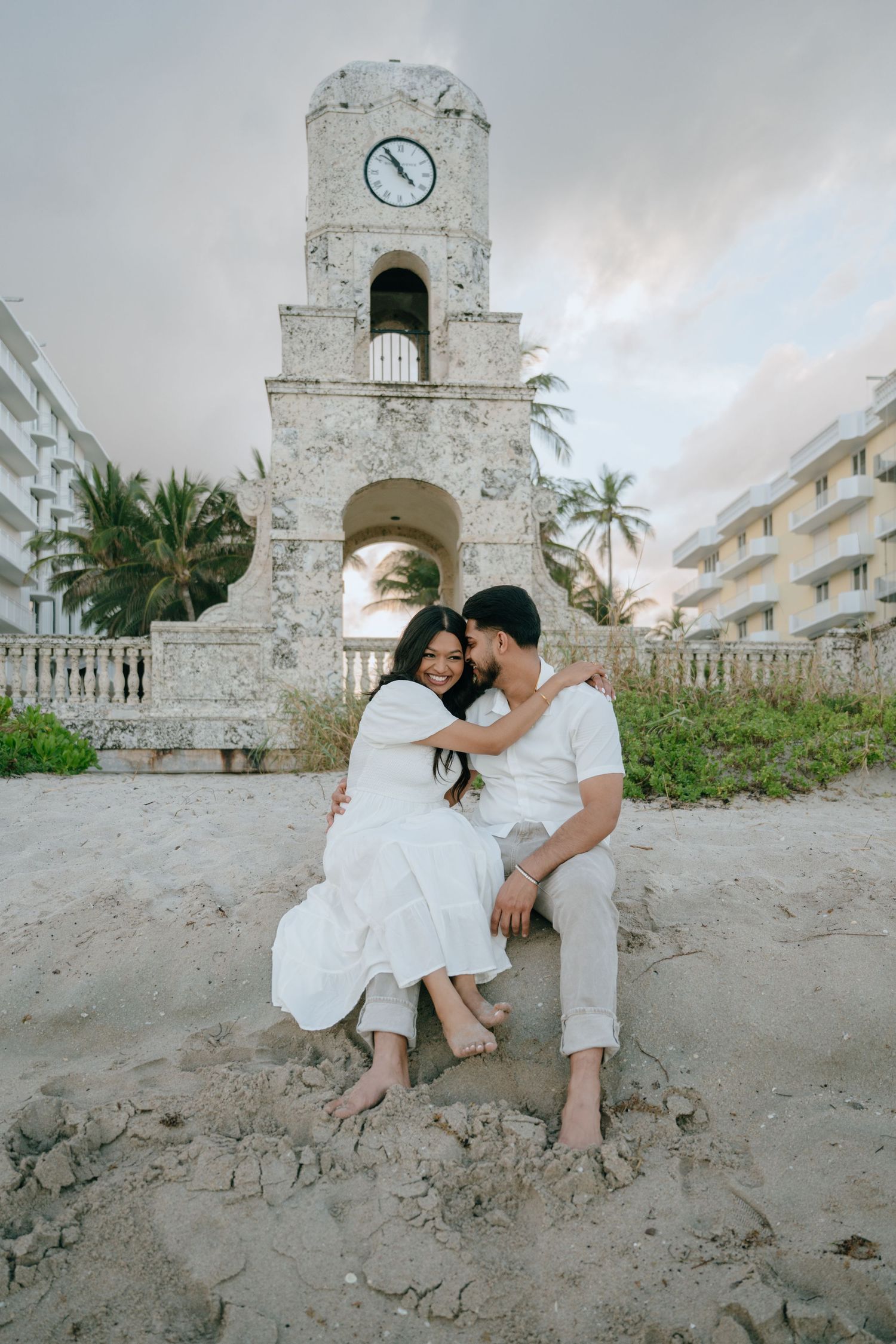 Couple in white outfits sitting together on sandy beach beside a vintage clock tower.