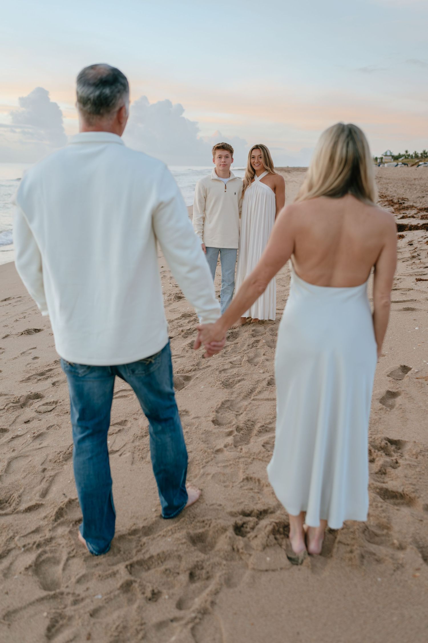 Parents hold hands facing their two teenage children on a beach at sunset.
