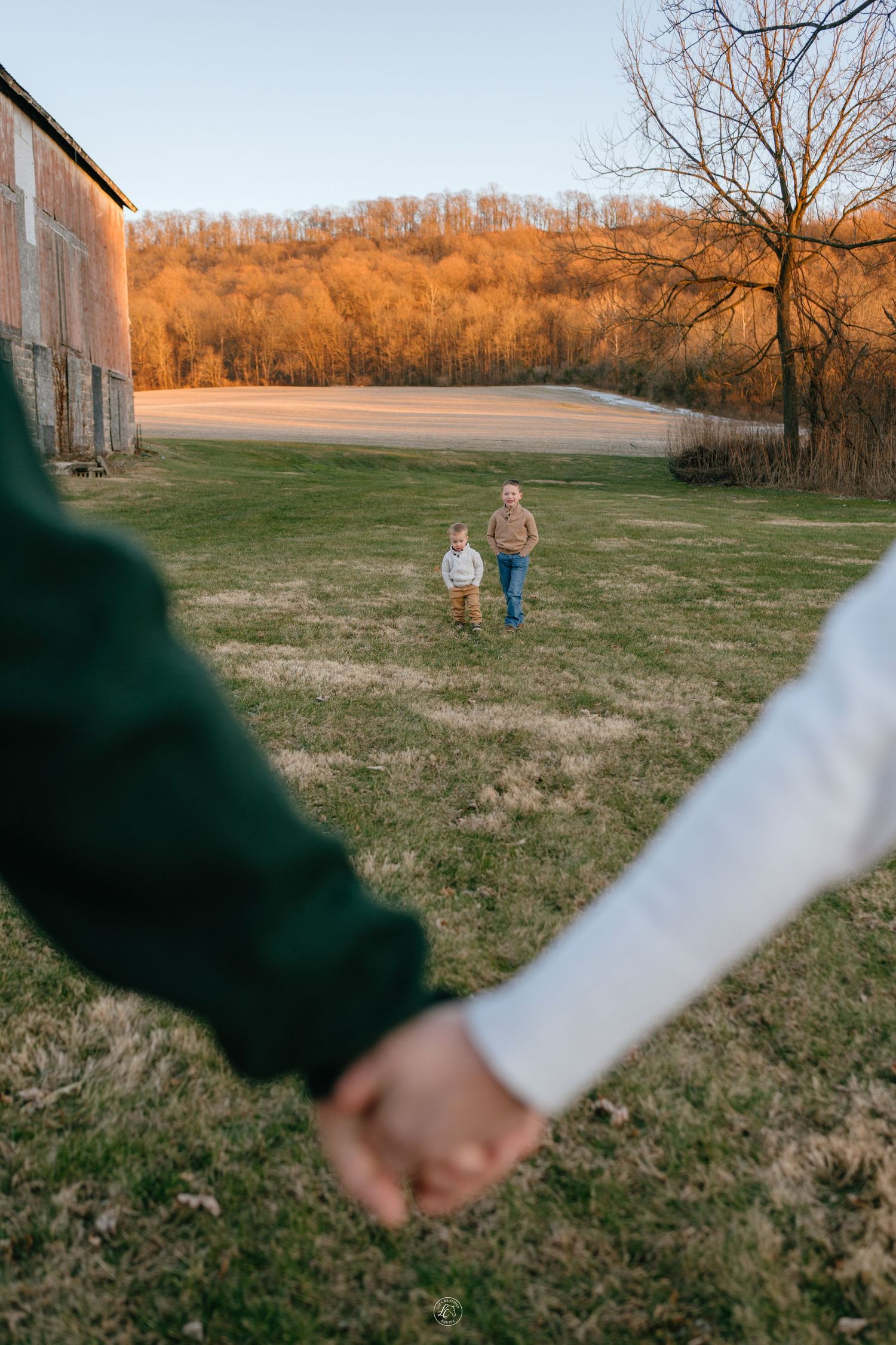 Parents holding hands in foreground while two young children run toward them on a farm at sunset.