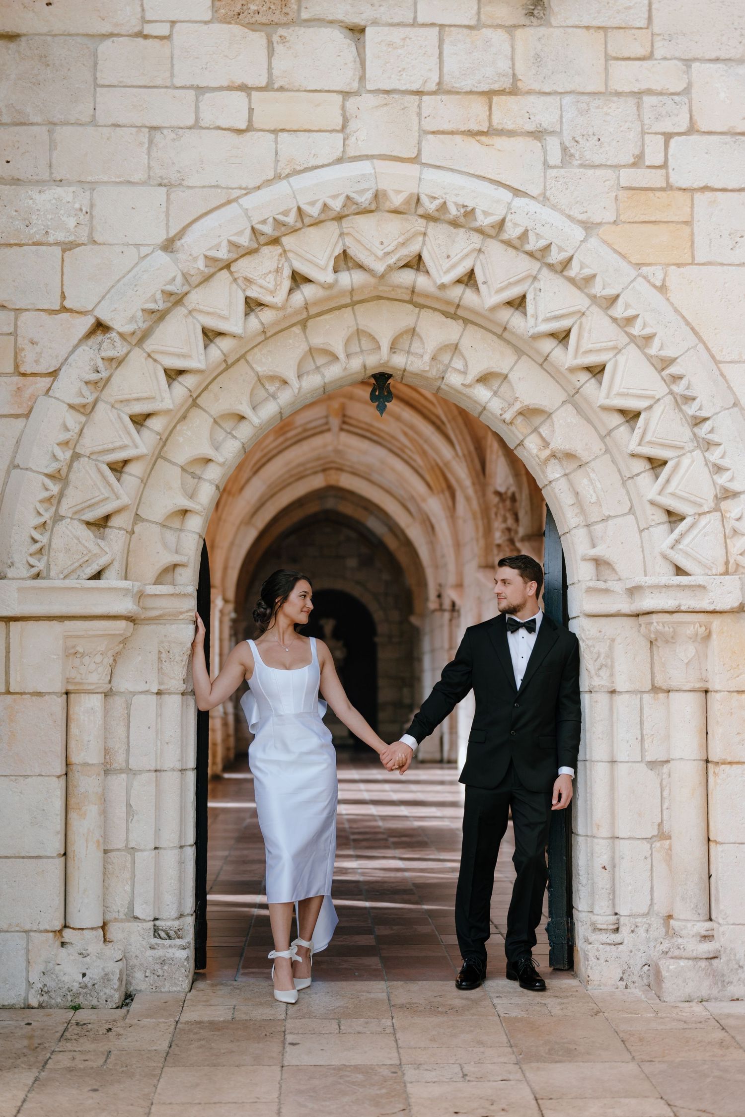 Bride and groom holding hands in elegant wedding attire beneath an ornate stone archway.