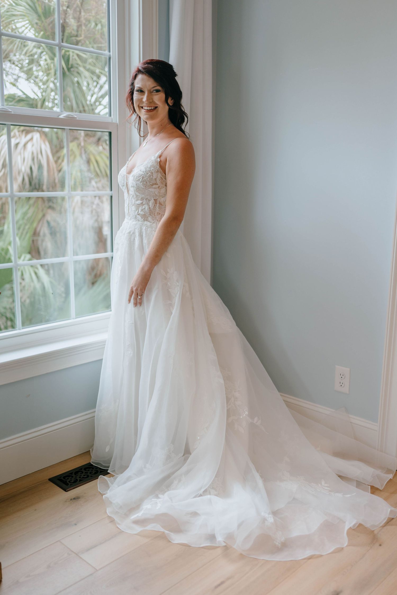 Smiling bride in lace A-line wedding gown with long train standing by a sunlit window.