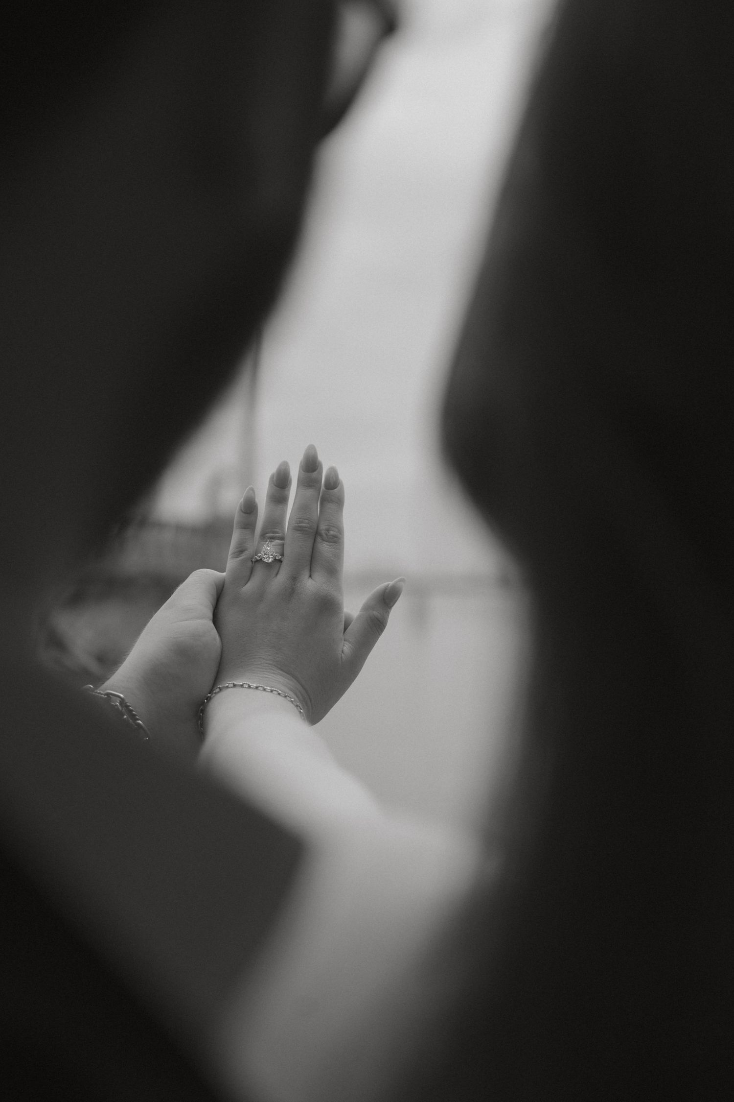 Black and white artistic close-up of hand displaying diamond engagement ring framed by couple silhouettes.