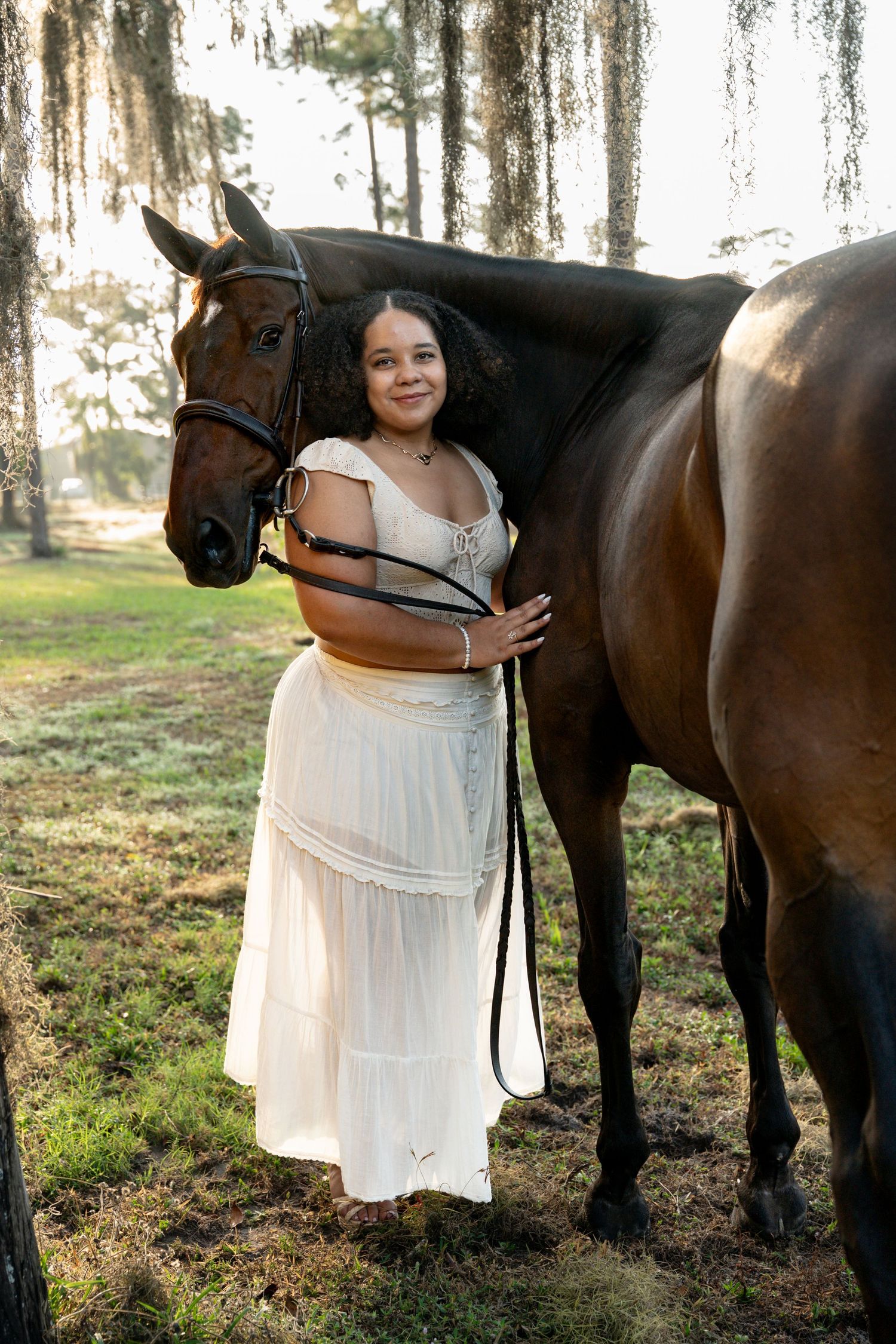 Smiling woman in white dress embraces dark bay horse in golden hour forest setting.