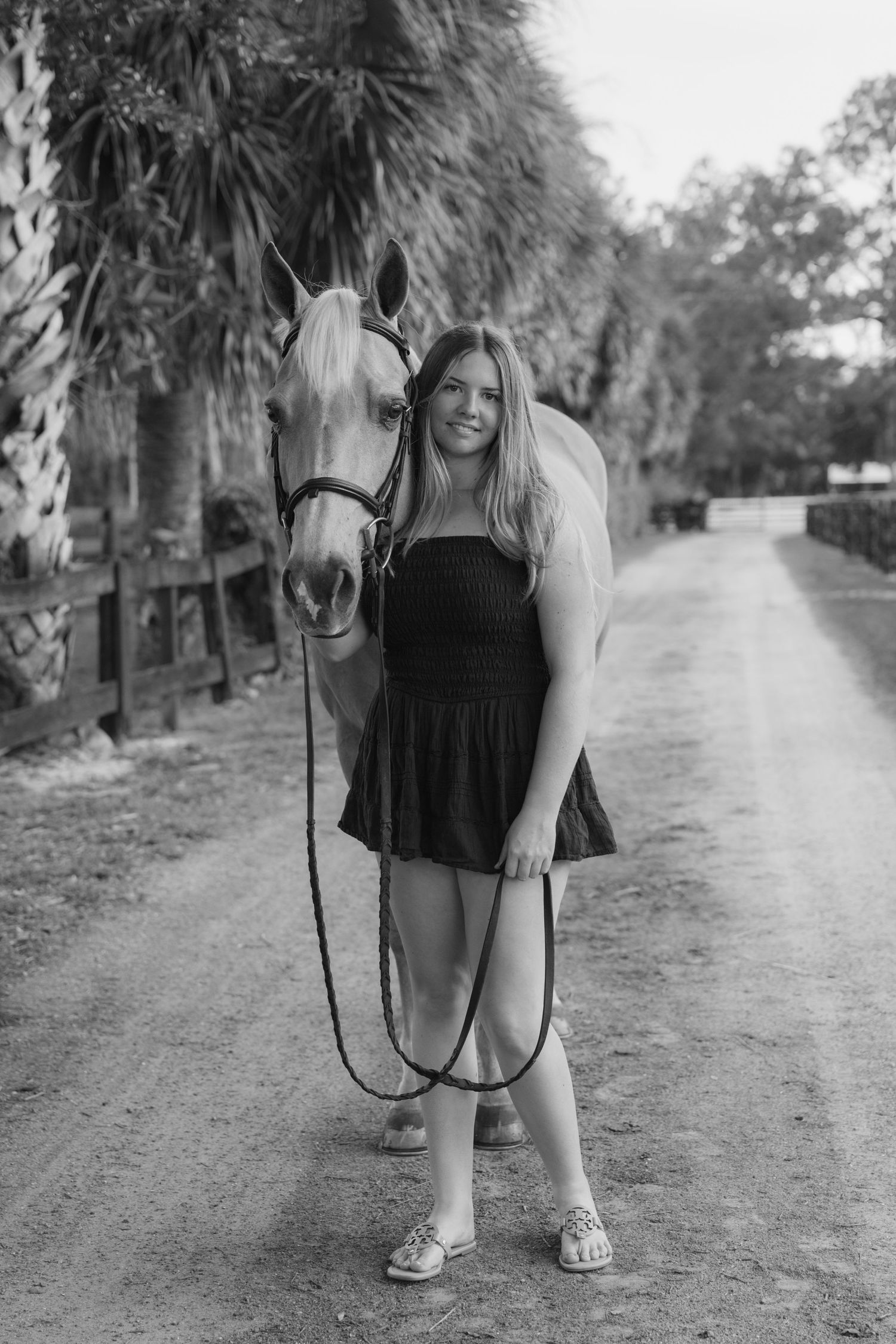 Young woman in black dress poses with a light-colored horse on a rural dirt path in black and white photo.