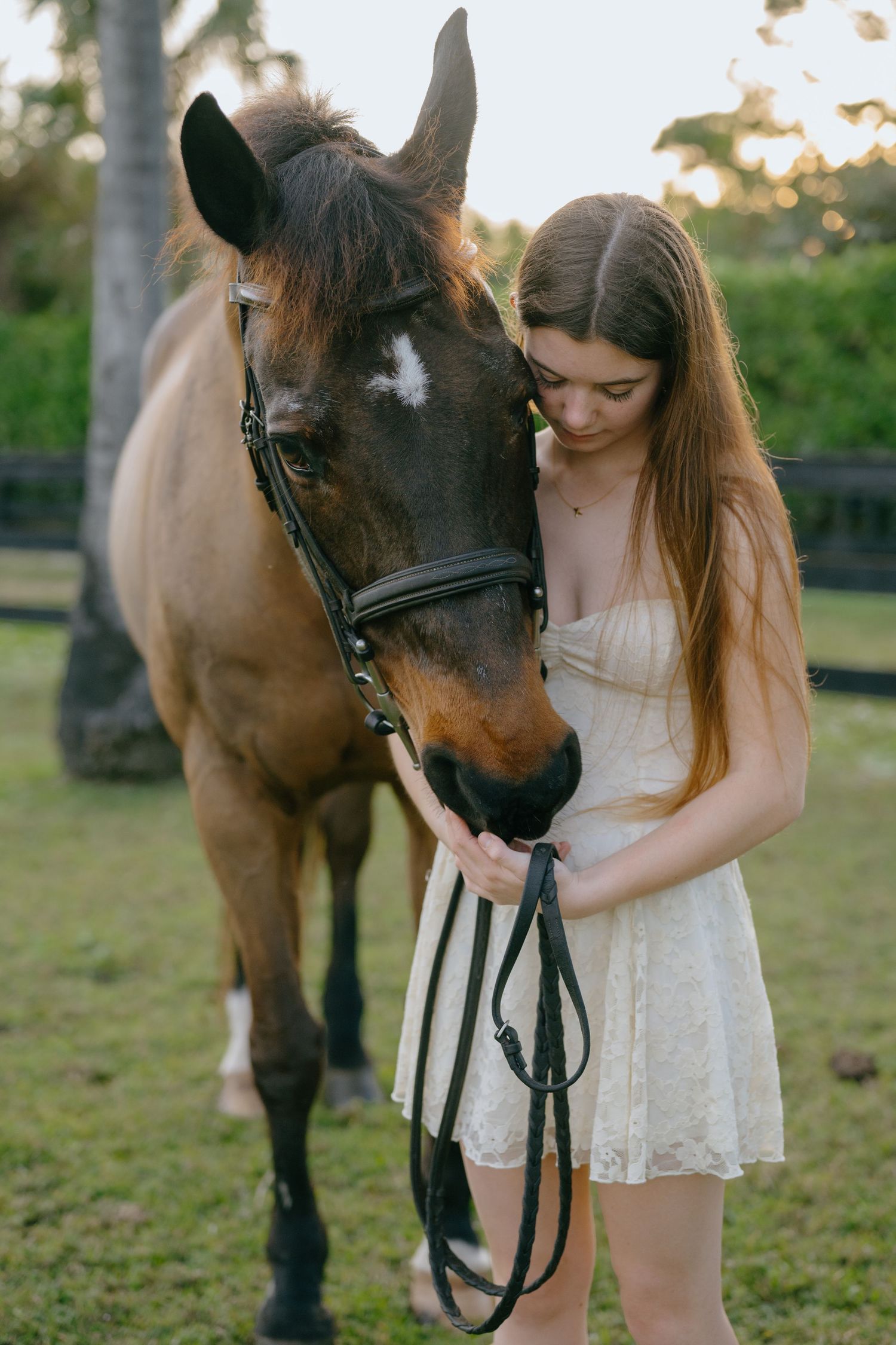 A young woman in a white dress tenderly holds the bridle of a bay horse in a green pasture.