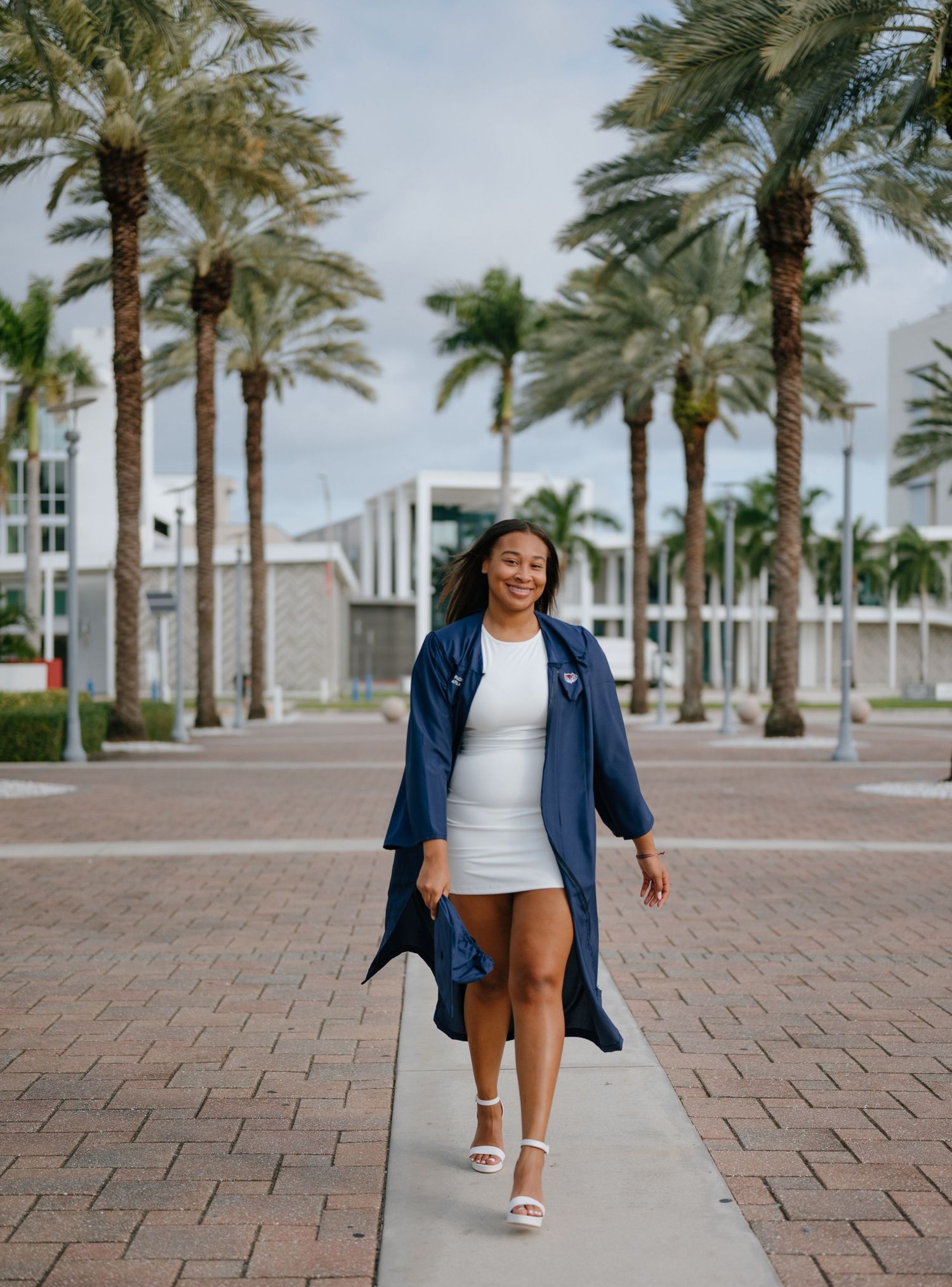 Young woman in navy graduation gown and white dress walks among palm trees at Florida Atlantic University.