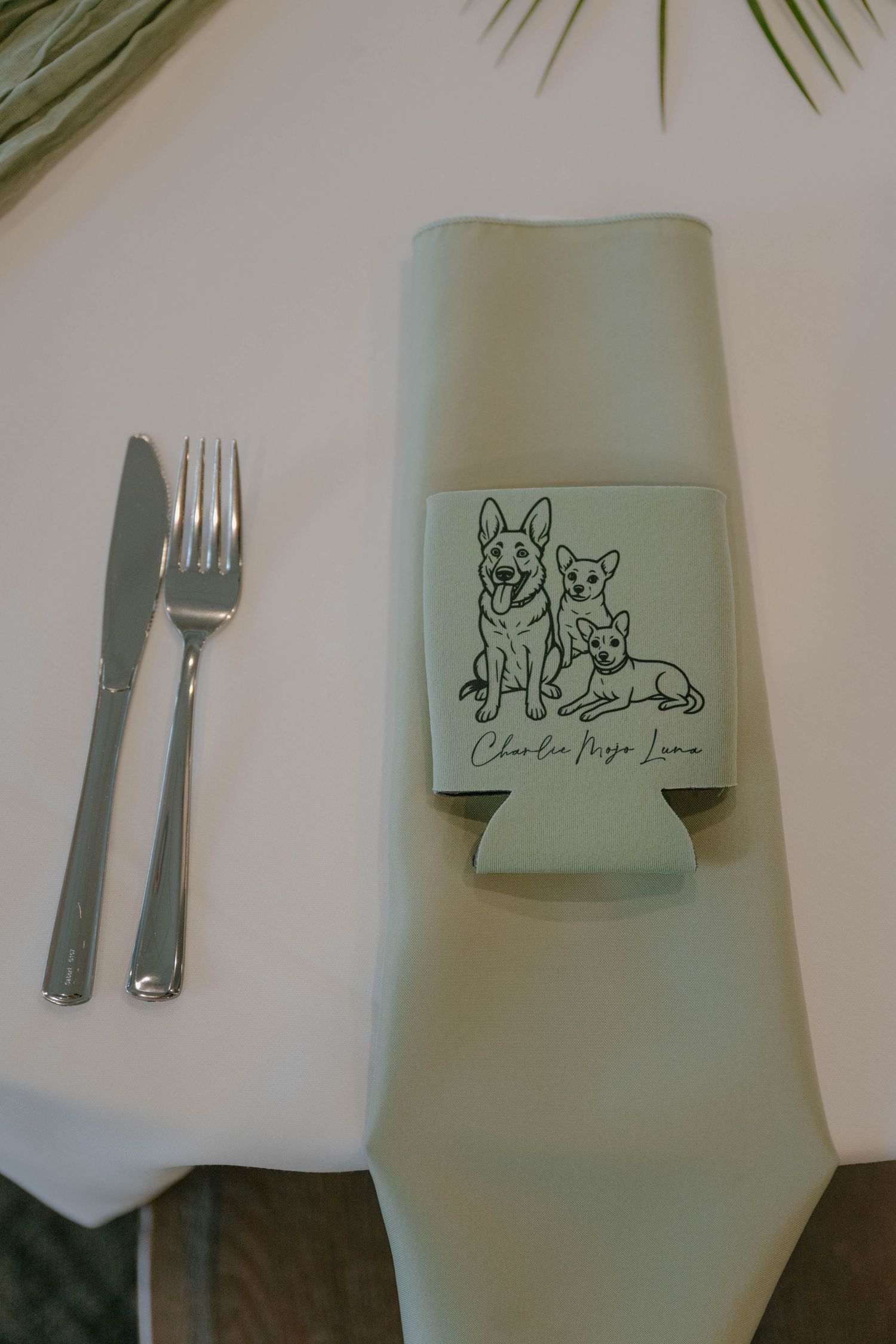 Custom wedding koozie with dog illustration on sage green napkin at elegant table place setting.