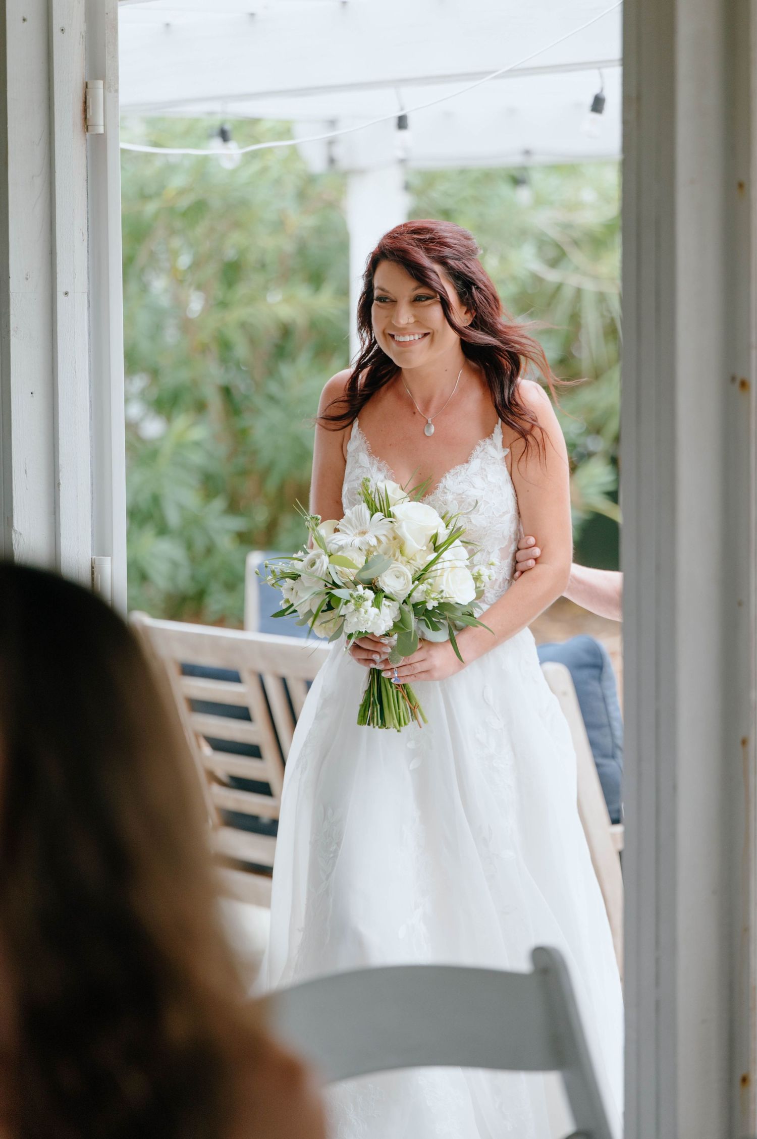 Smiling bride in white lace gown holding white floral bouquet standing in doorway on wedding day.